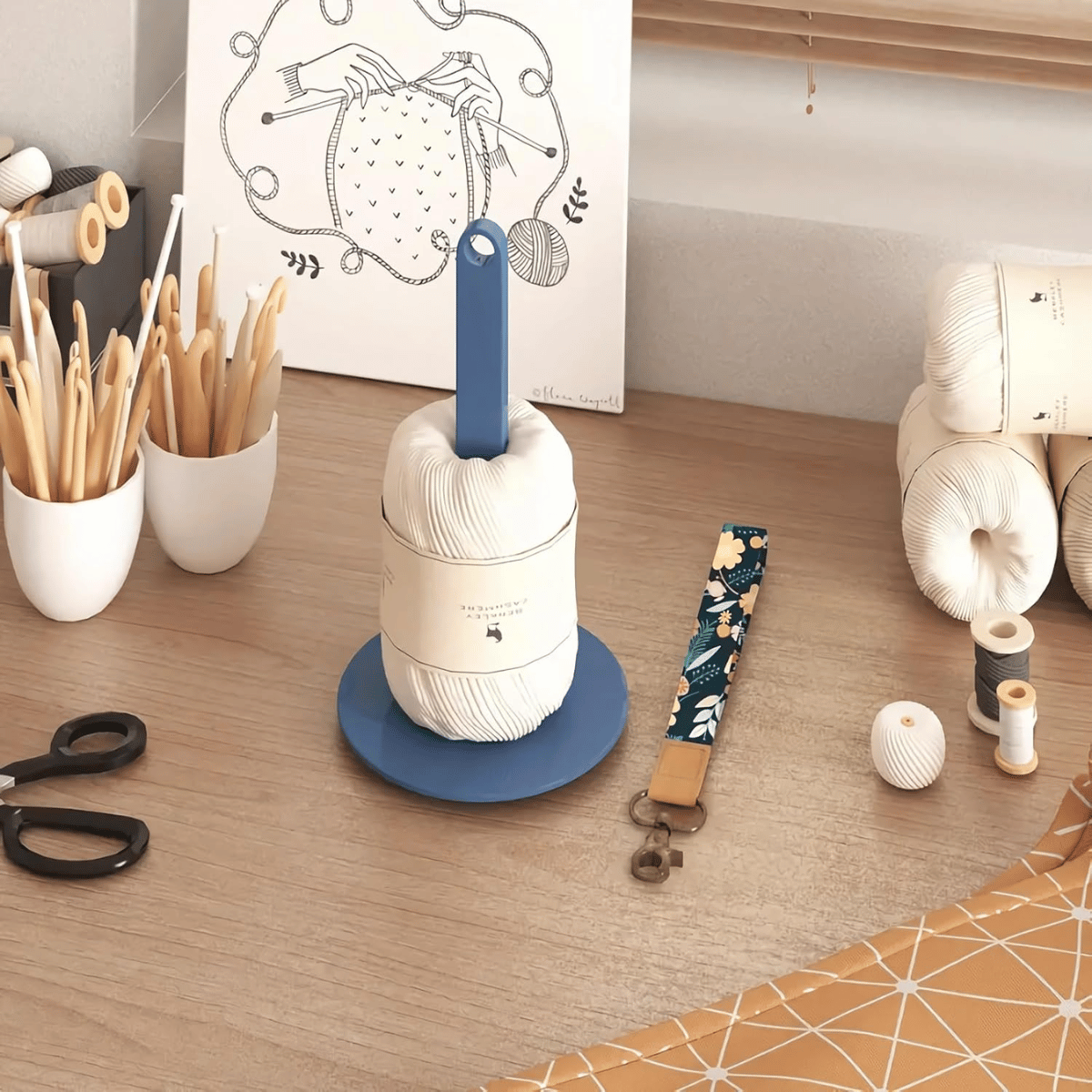 A tidy craft workspace displays the Wrist Yarn Holder for hands-free yarn control, crochet hooks in white cups, scissors, a floral lanyard, and an illustrated knitting print—perfect for crafting enthusiasts on the go.