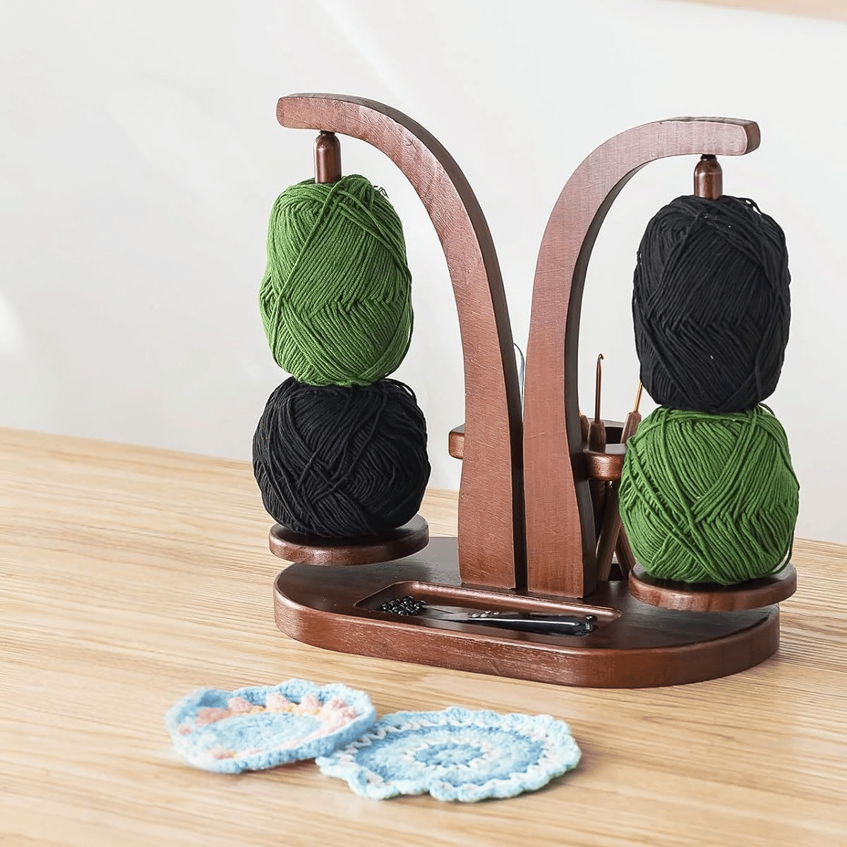 The Double Yarn Spinner Stand with Tool Storage keeps yarn tangle-free and organized, holding four balls—two green, two black—next to blue, white, and pink crocheted coasters.