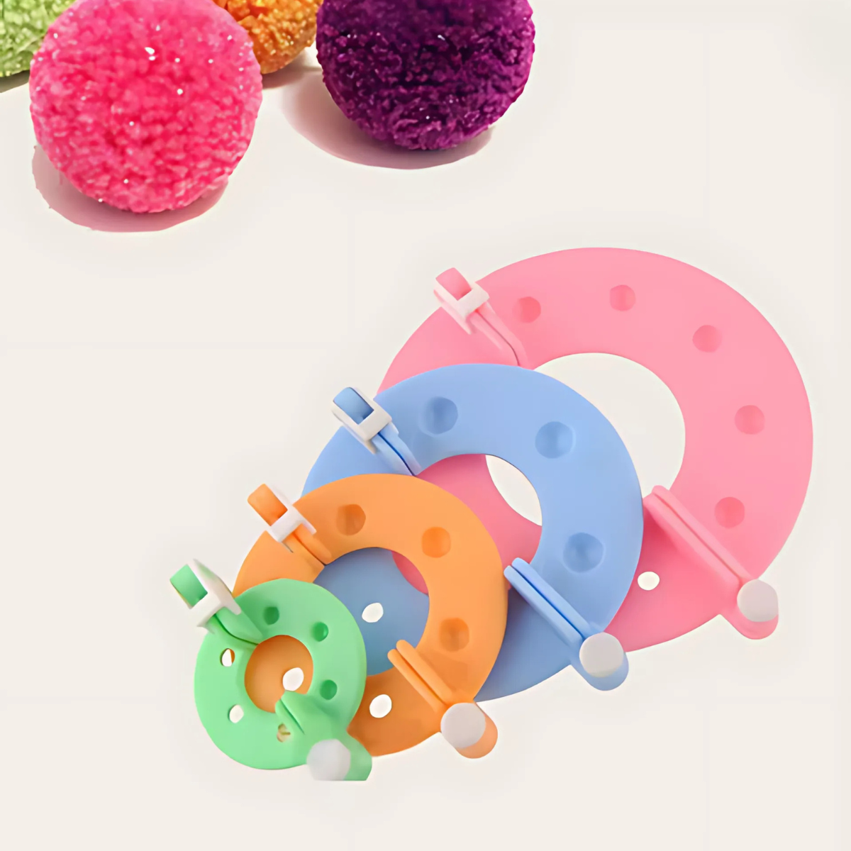 The Pom Pom Maker Set includes four plastic tools in green, yellow, blue, and pink, arranged by size, with three pom poms in the background—ideal for crafting projects.