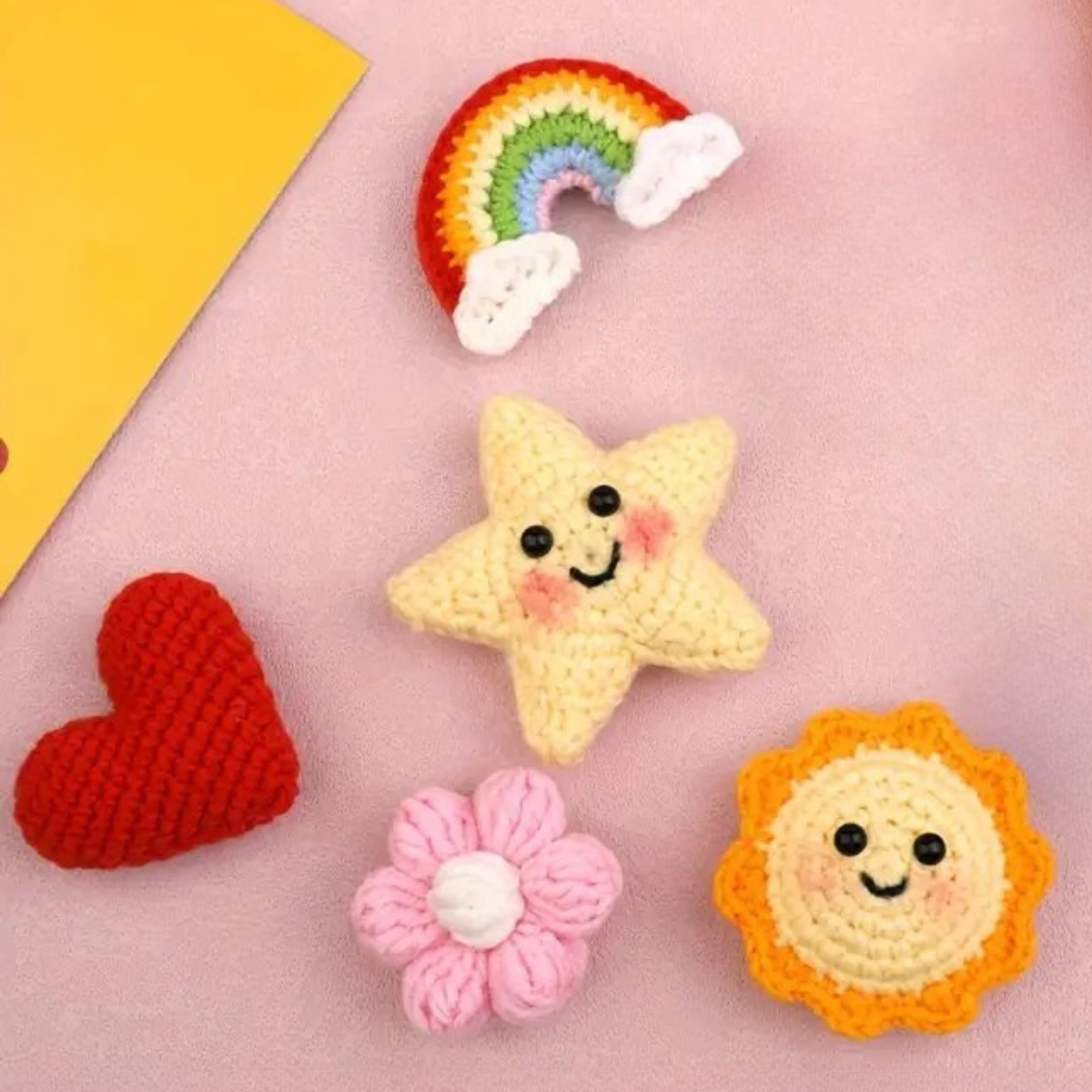 Four crocheted items—red heart, rainbow, pink flower, and yellow smiley sun—are displayed on a light pink surface, perfect for making your own handmade keychains with our Crochet Keychain Kit 4pcs.