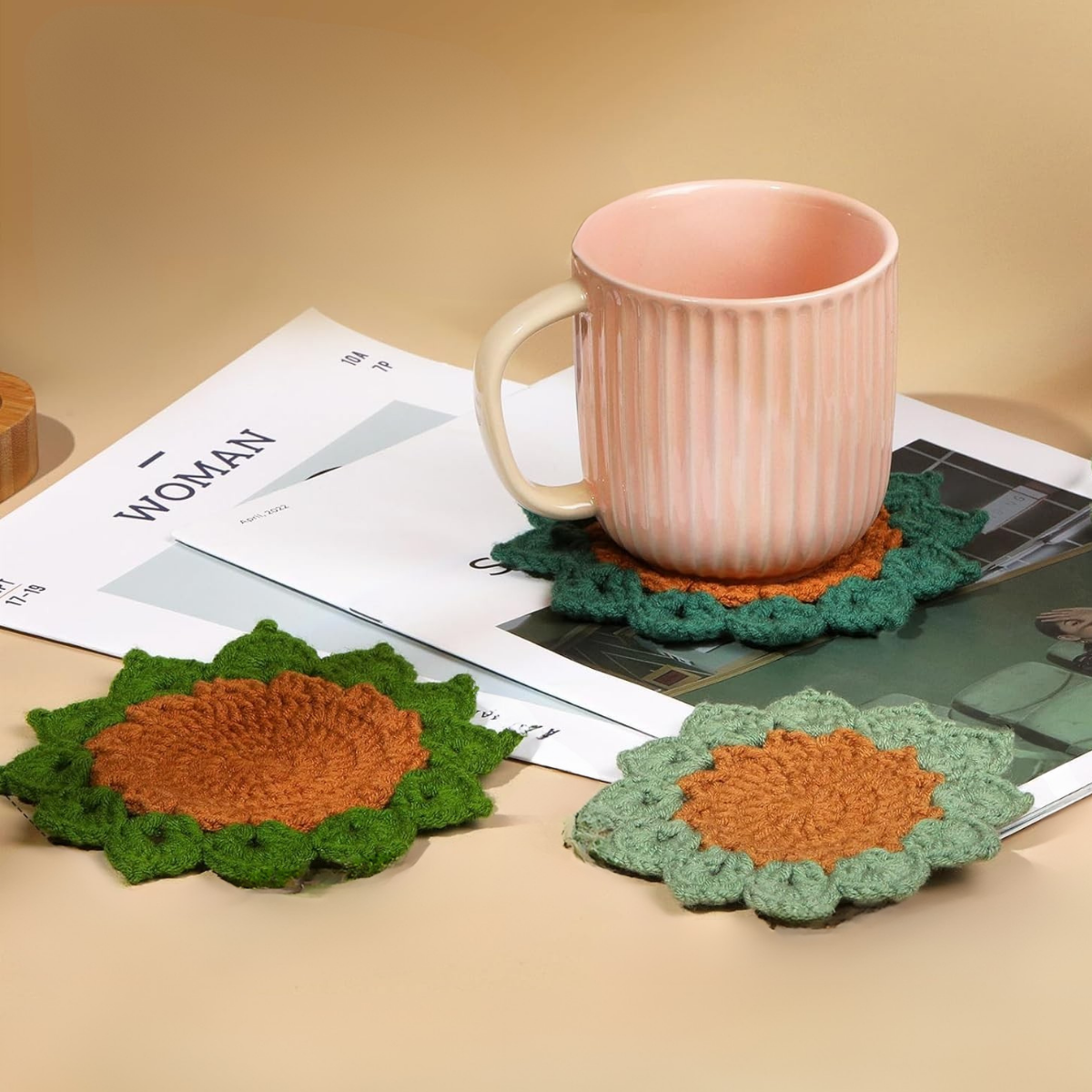 A pink ceramic mug sits on a green and brown coaster made from the Plant Pot Crochet Kit with Coasters, with two matching coasters and magazines nearby on a beige surface.