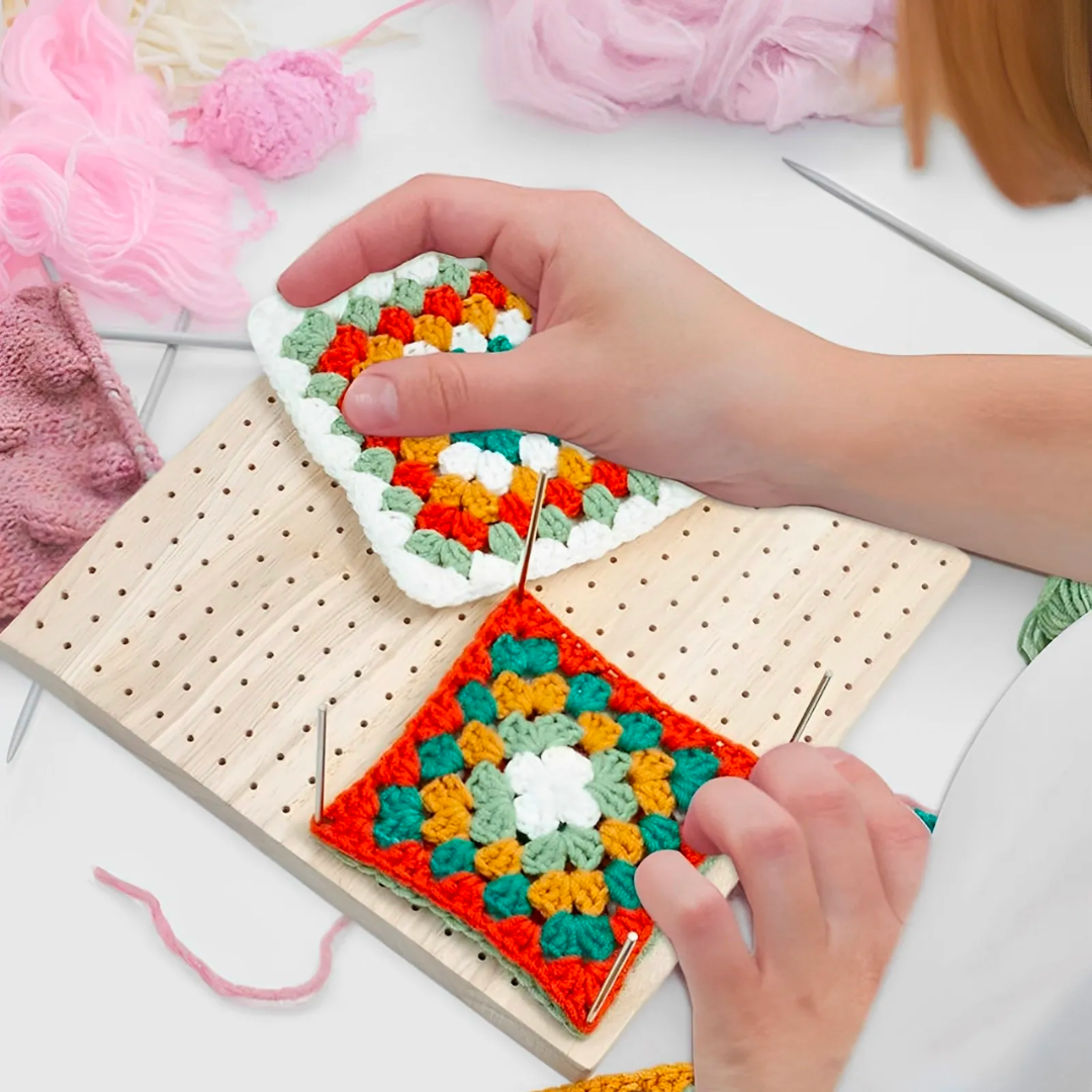 Hands carefully arrange vibrant crocheted squares on a Crochet Blocking Board, surrounded by crochet tools and yarns, highlighting the meticulous artistry of a dedicated crocheter.