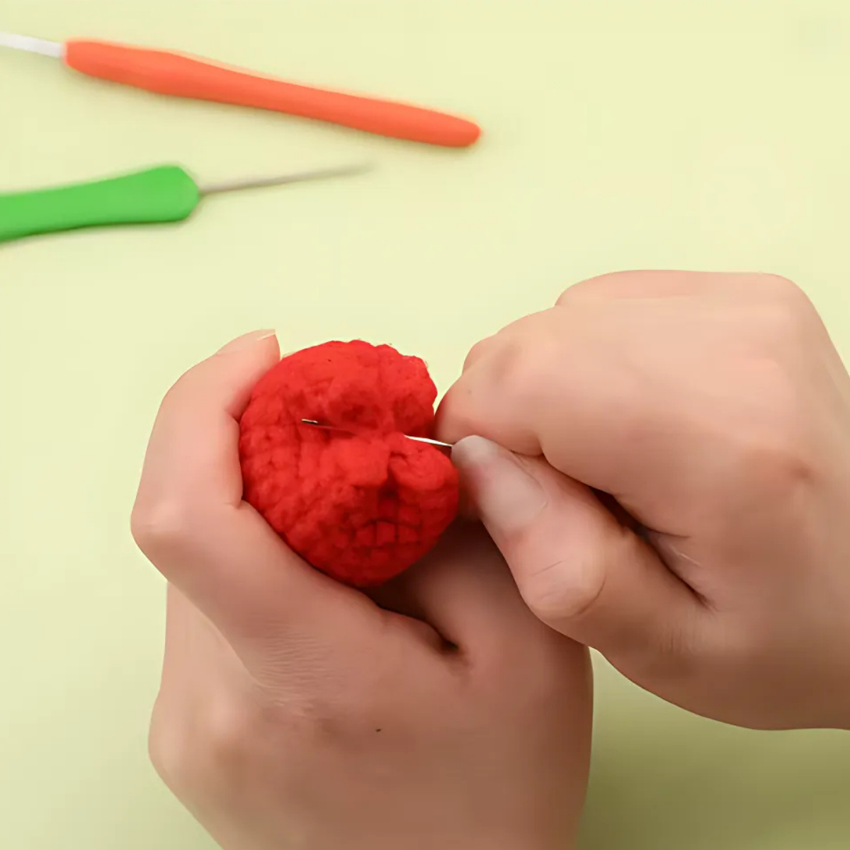 Two hands sew a red crocheted piece with a needle, while green and orange crochet hooks rest nearby—an ideal scene for using the Crochet Flower Pot Kit 6pcs, great for beginners making handmade crochet flowers.