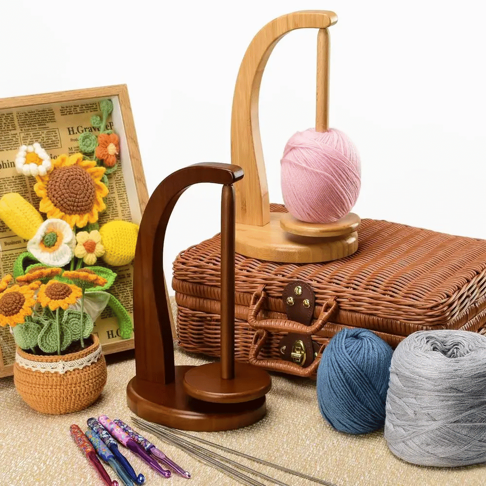 Two Levitating Premium Yarn Holders with balls of yarn promise a seamless knitting experience. Nearby, a wicker basket cradles crocheted sunflowers in a pot, while crochet hooks rest on the textured surface.