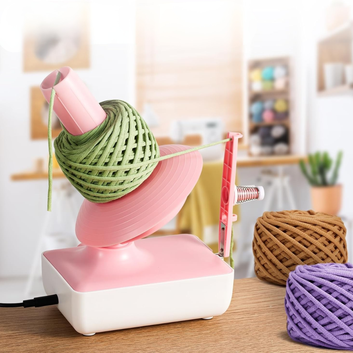 An Electric Yarn Winder in pink and white sits on a table, winding green yarn, with brown and purple yarn balls nearby in a bright, modern room.