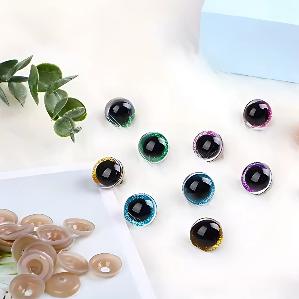 The Safety Eyes & Noses Set features vibrant crochet eyes with black centers, displayed on a white surface near green leaves, beige buttons, and a light blue box—ideal amigurumi supplies for your next project.