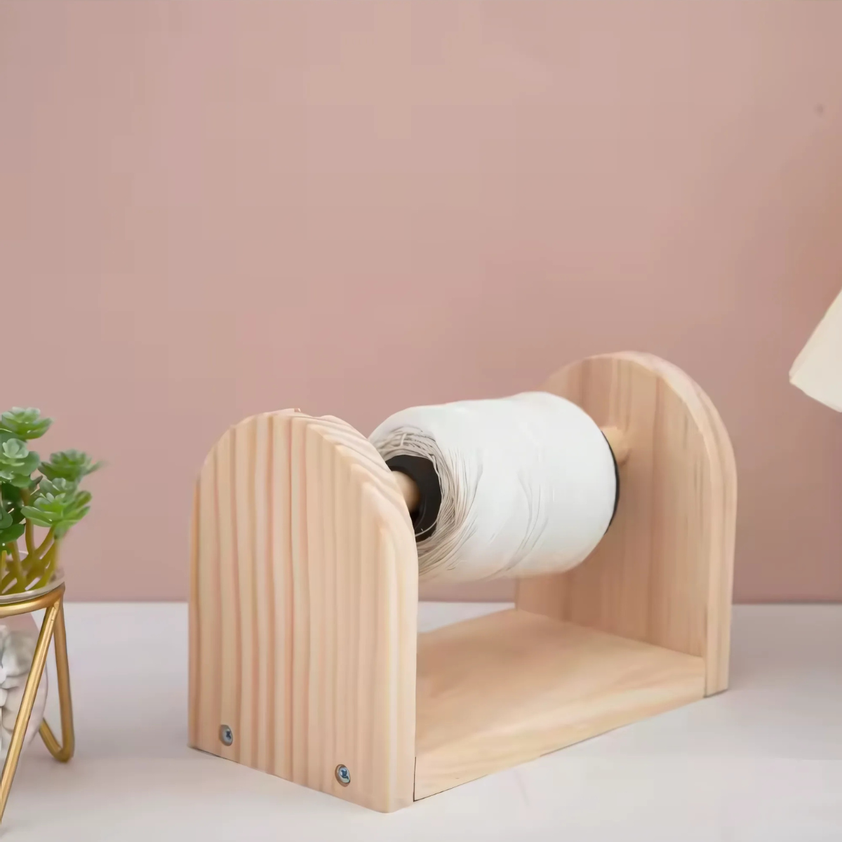 The Wooden Yarn Dispenser, shown on a white surface beside a potted plant, securely holds and smoothly rotates a paper towel roll—ideal for crafting or daily use.