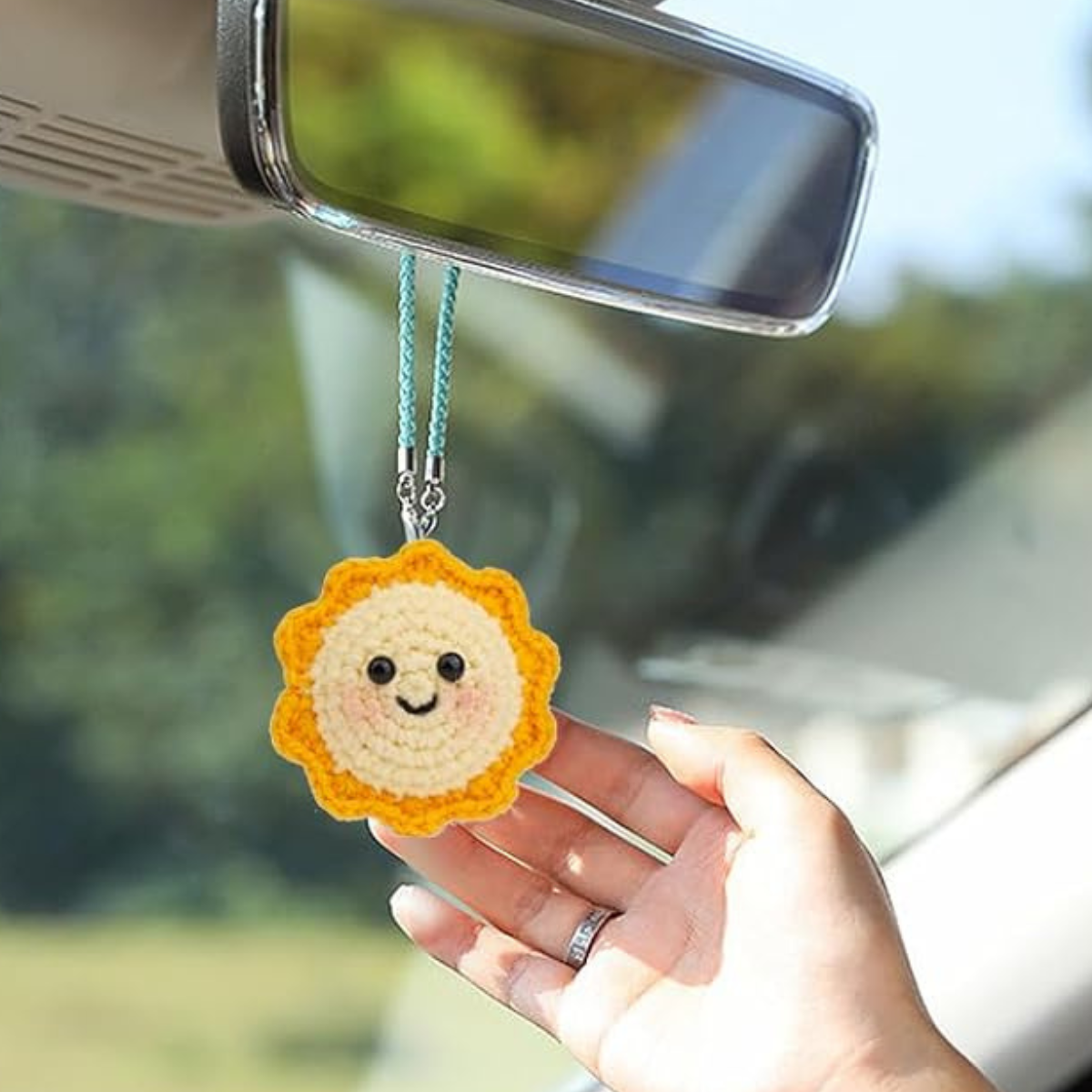 A hand holds a sun smiley charm from the Crochet Keychain Kit 4pcs, handmade and hanging by a blue strap from a car’s rearview mirror.