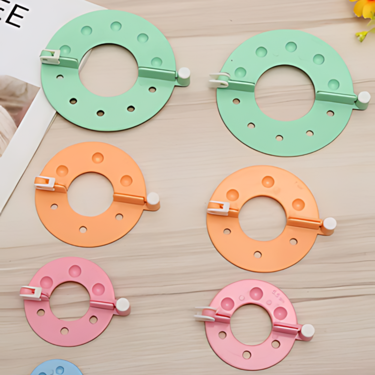 The Pom Pom Maker Set includes six circular plastic tools in green, orange, and pink with white sliding latches and holes, arranged on a wooden surface—perfect for crafting pom poms.