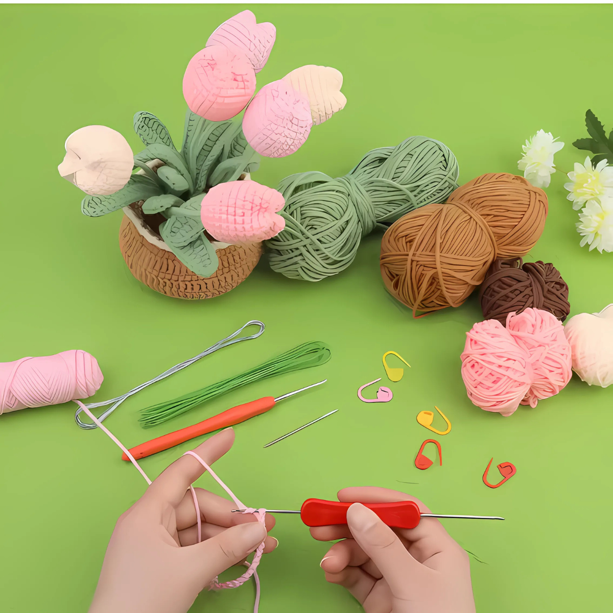 Two hands crochet with a red hook, surrounded by yarn, tools, and stitch markers beside a basket of yarn flowers—perfect for using the Crochet Tulip Kit to create your own beginner crochet flower bouquet—set on a green background.