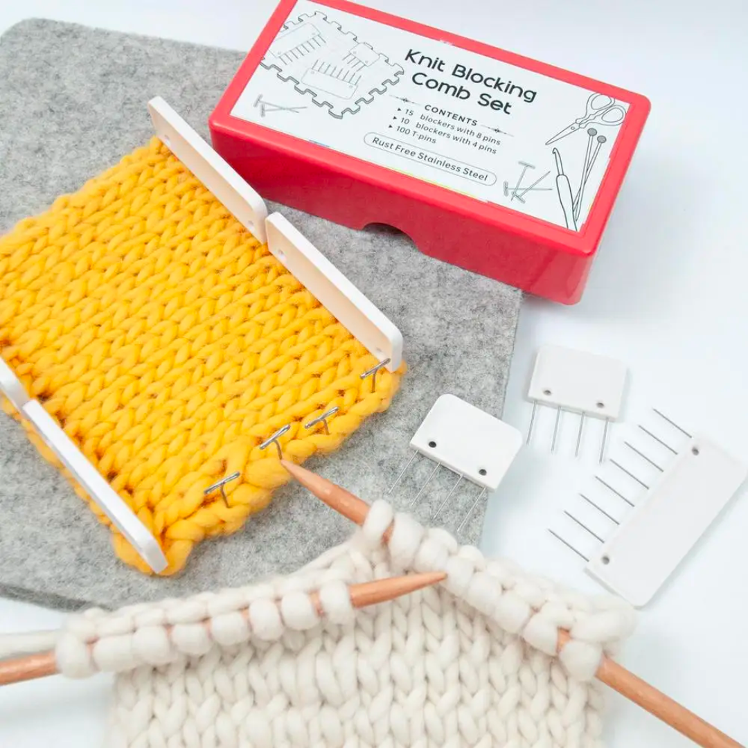 The Knit Blocking Comb Set features durable blocking pins and is packaged in a red box adorned with a yellow knit swatch on a gray mat. It includes stainless steel comb pieces and a white swatch accompanied by knitting needles, ensuring perfect alignment for your projects.