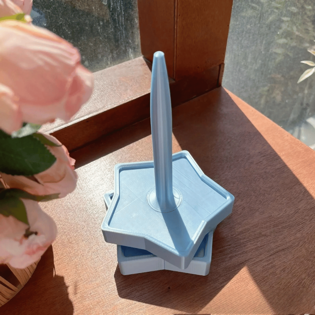 A Spinning Yarn Holder sits on a wooden surface next to a flower arrangement by a sunlit window.