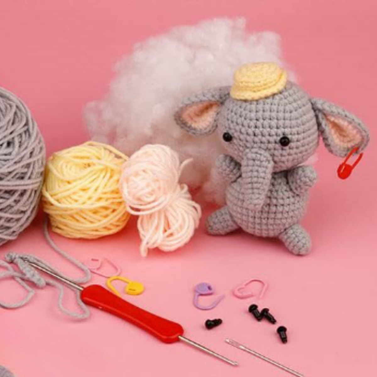 The Crochet Elephant Kit includes yarn balls, stuffing, crochet hooks, plastic markers, and toy eyes—all arranged with a crocheted elephant plush on a pink background.