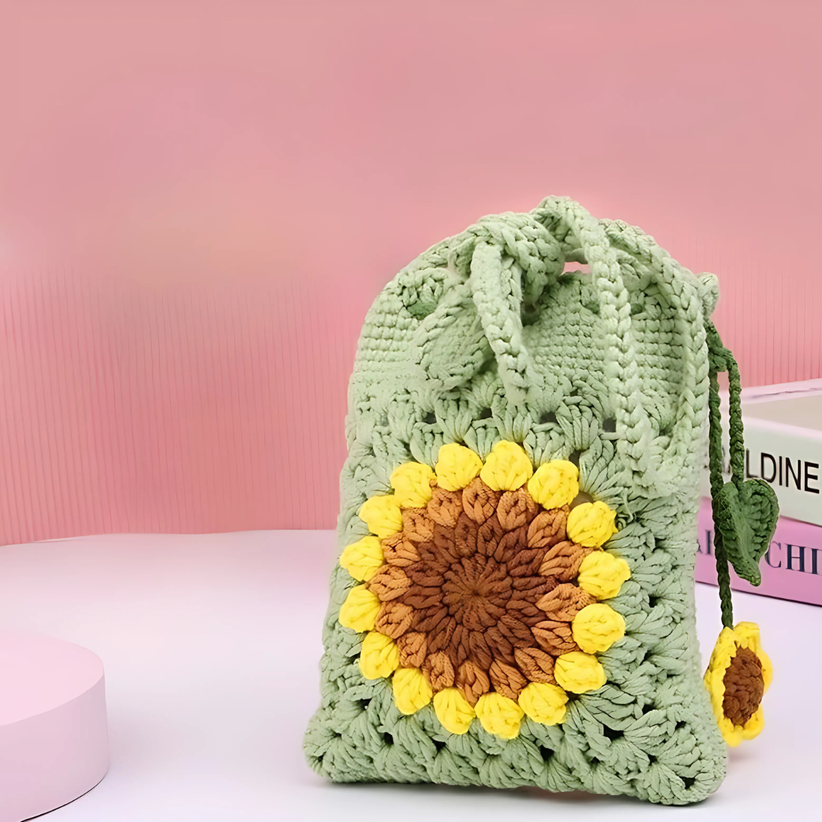 A green Crochet Bag Kit featuring a large crocheted sunflower design, shown against a pink background with books nearby—ideal for anyone wanting a unique accessory or to craft their own sunflower-inspired bag.