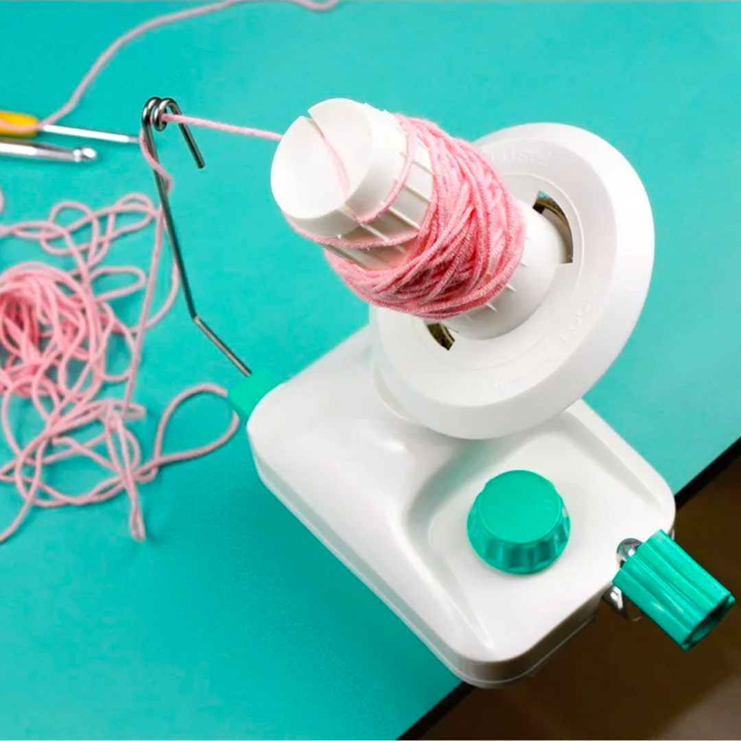 A Yarn Ball Winder is secured to the edge of a turquoise table, accompanied by pink yarn, making it an ideal setup for your crocheting projects. Nearby, loose yarn balls await your next creation.