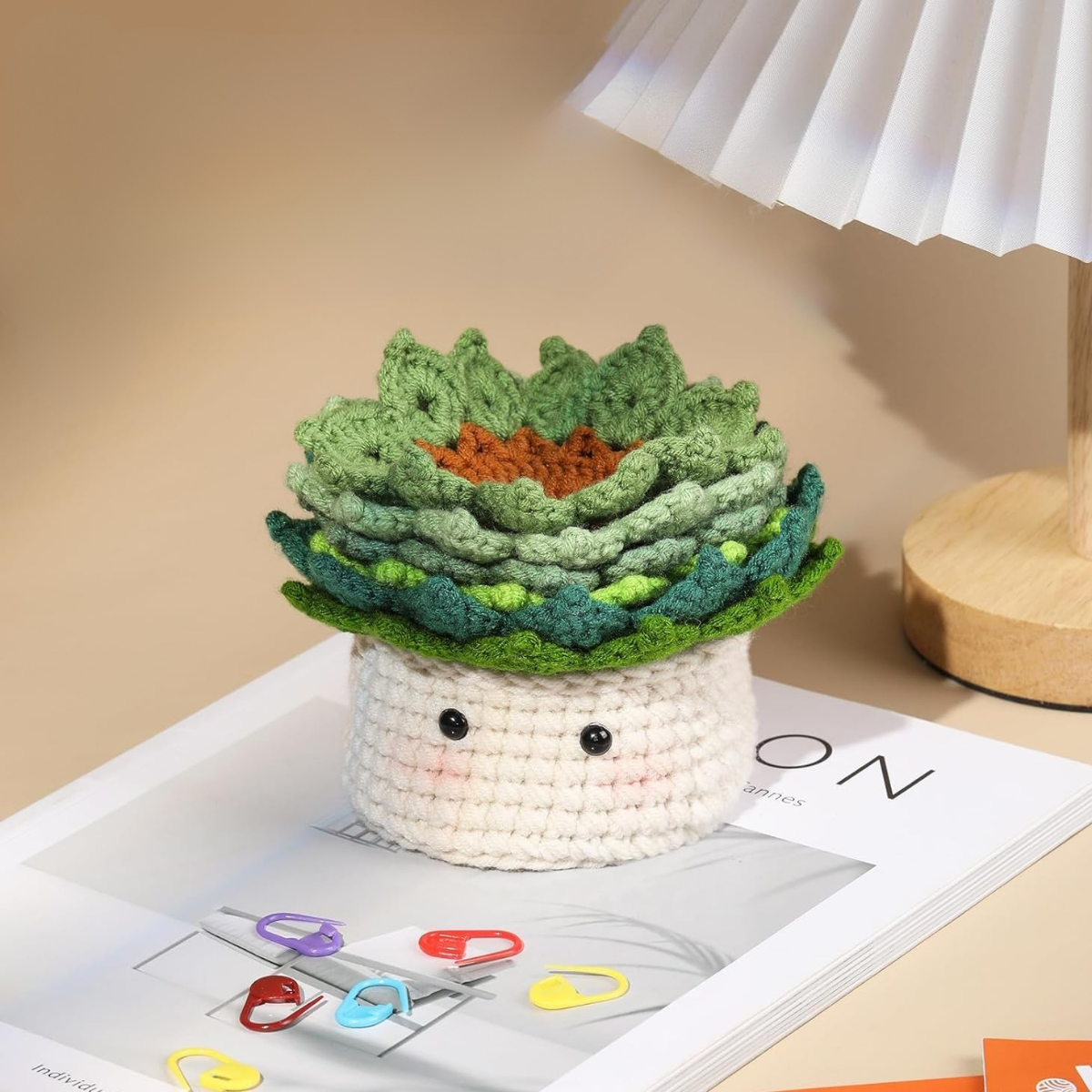 The Plant Pot Crochet Kit with Coasters features a crocheted succulent in a white base, displayed on a magazine beside colored paper clips and a lamp for charming handmade home decor.