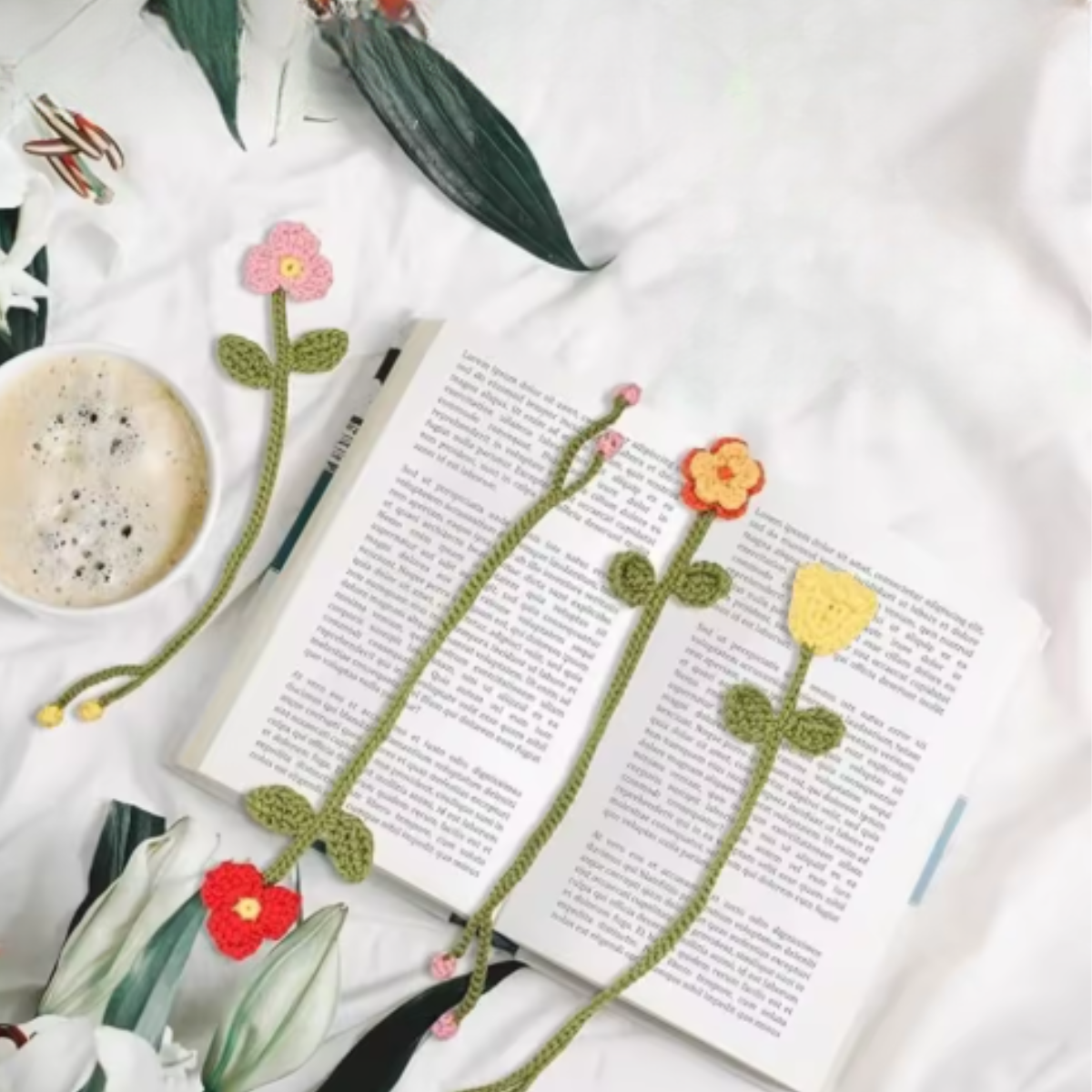 Eight handmade bookmarks from the Crochet Flower Bookmark Kit, each featuring a crocheted flower, rest on an open book with coffee and green leaves—ideal gifts for book lovers.