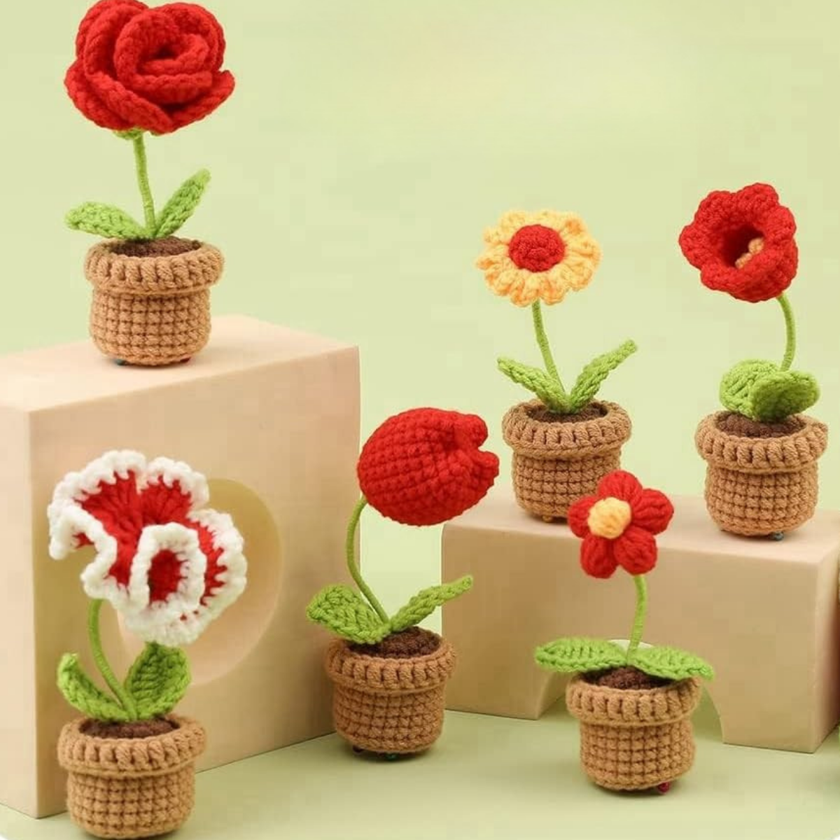 The Crochet Flower Pot Kit 6pcs features six small handmade crochet flowers in assorted colors and shapes, beautifully displayed on light wooden blocks against a pale green background.