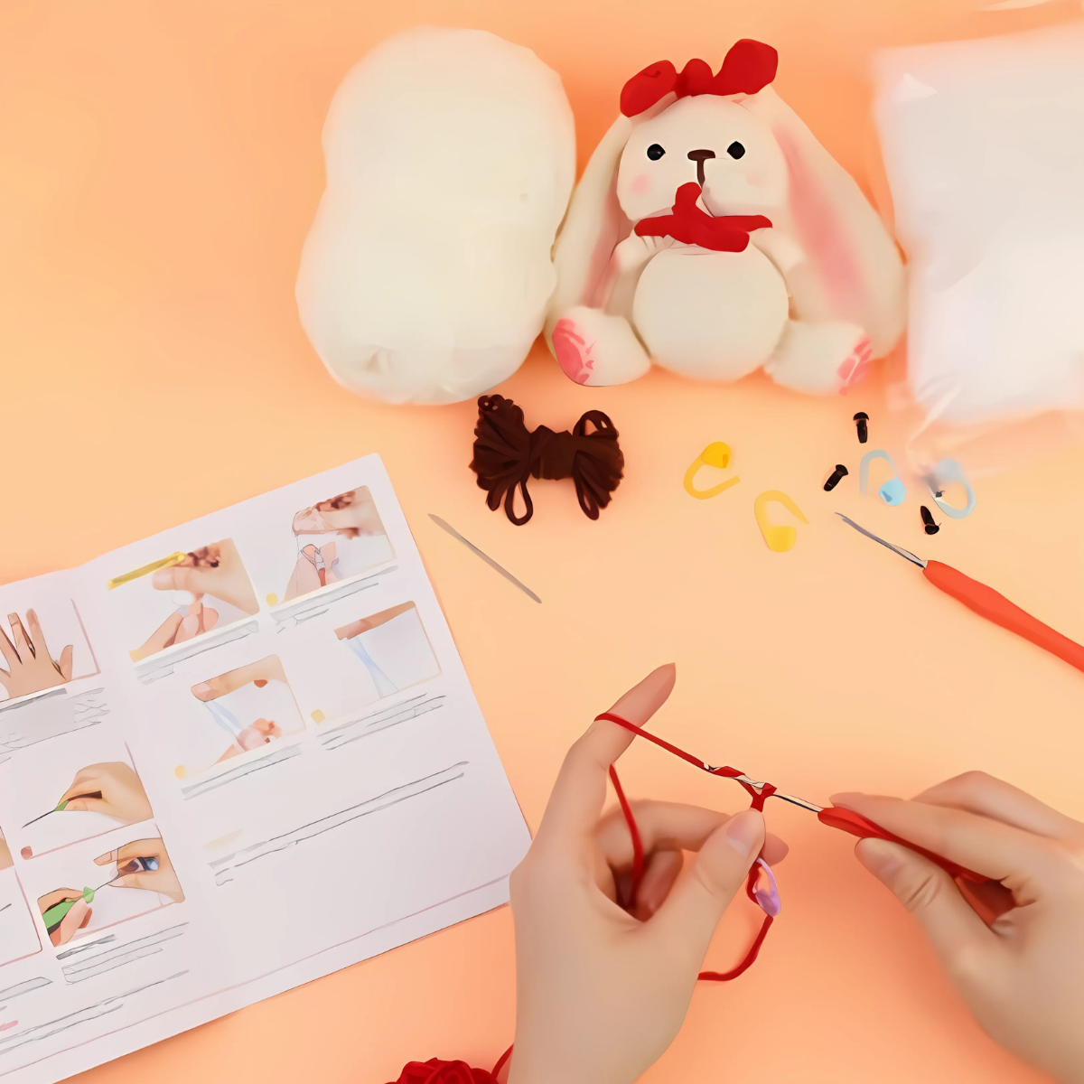 Hands crocheting with a red hook, surrounded by yarn, crochet tools, a partially made bunny, and illustrated instructions—everything included in the Crochet Rabbit Kit for beginners on a peach-colored surface.