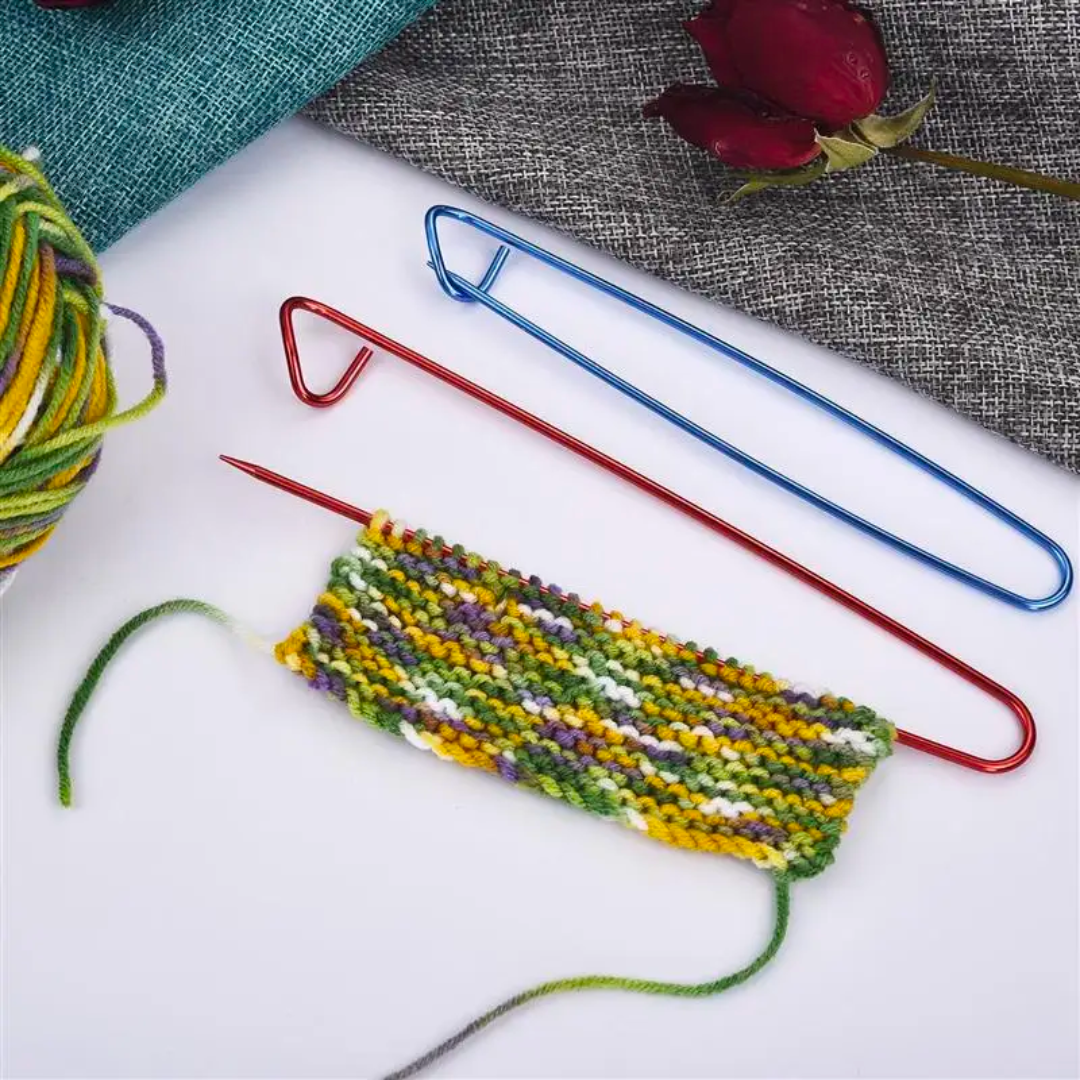 A vibrant knitting project features a small striped piece on a red needle, enhanced by the durable Stitch Holder Set 6pcs. Nearby rests a ball of multicolored yarn and a delicate red rose on gray fabric, creating an inviting scene for any knitting tools enthusiast.