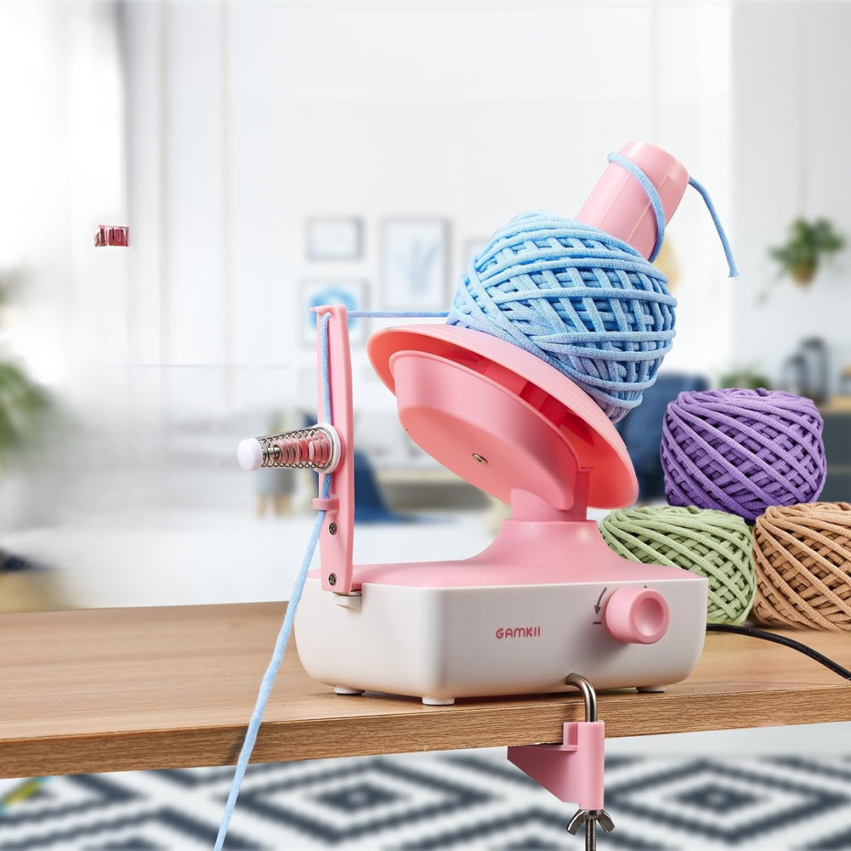 An Electric Yarn Winder securely clamped to a table quickly winds blue yarn into a tidy ball, while more colorful yarn balls sit nearby.