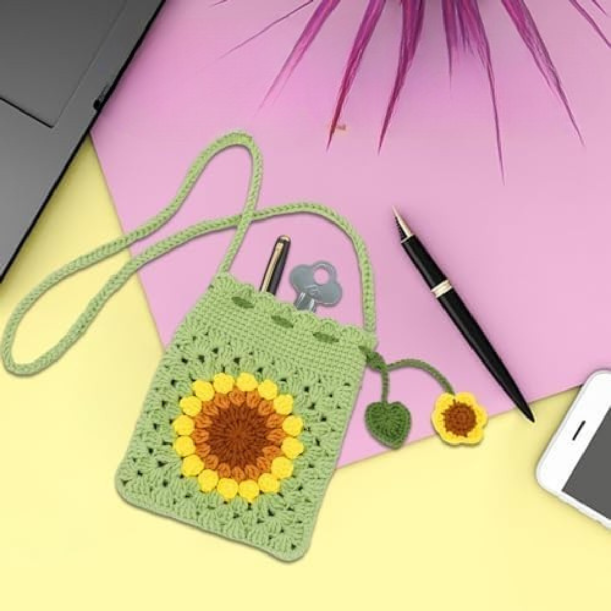 A green sunflower bag made with the Crochet Bag Kit holds a pen, key, and pencil, and sits on a pastel yellow and pink desk next to a pen, laptop, and smartphone.