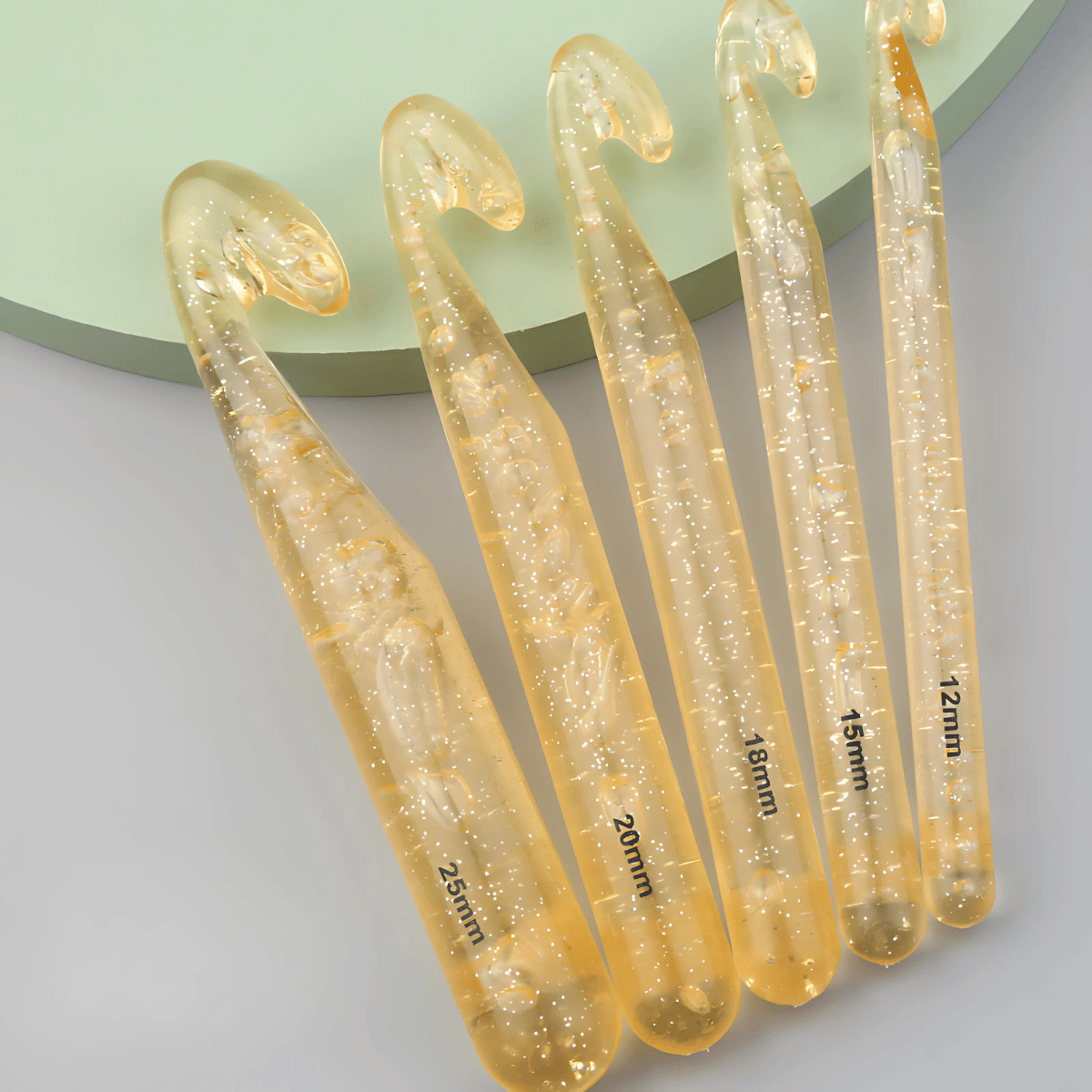 The Large Crochet Hooks for Chunky Yarn set includes five lightweight translucent gold glitter hooks (25mm, 20mm, 18mm, 16mm, and 12mm), perfect for jumbo yarn projects on a light green and gray background.