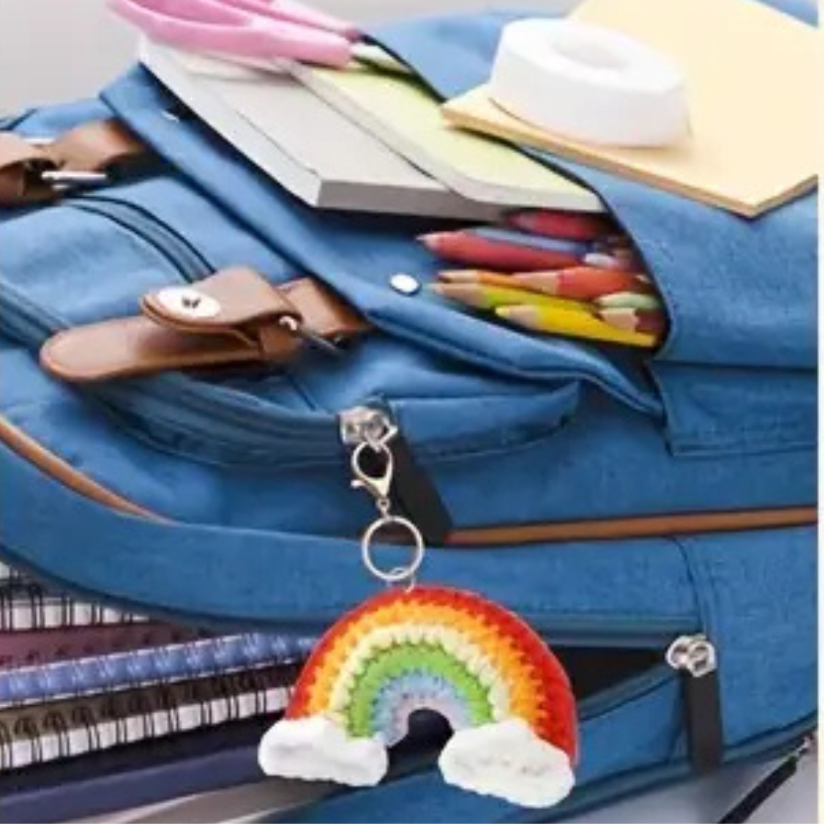 A blue backpack with colored pencils, notebooks, and stationery, featuring a keychain from the Crochet Keychain Kit 4pcs attached to the zipper.