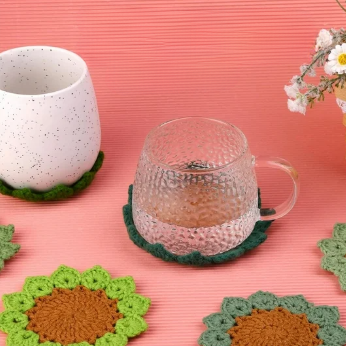 The Plant Pot Crochet Kit with Coasters features green crochet coasters displayed with a clear glass cup, a white ceramic mug, sunflower-patterned accents, and small white flowers on a cheerful pink background.
