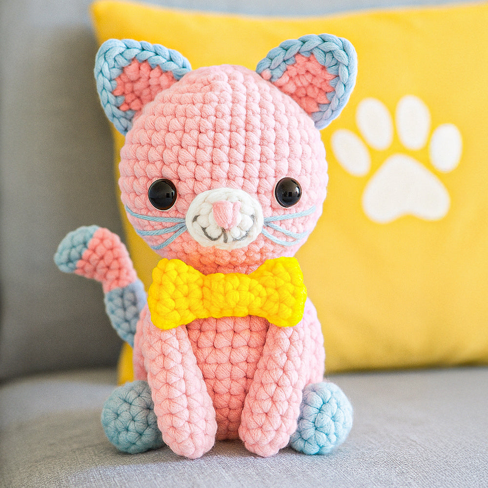Luna The Cat, a pink amigurumi plush with blue ears, paws, and tail tip and a yellow bow, sits on a couch beside a yellow pillow with a white paw print—a delightful intermediate crochet creation.
