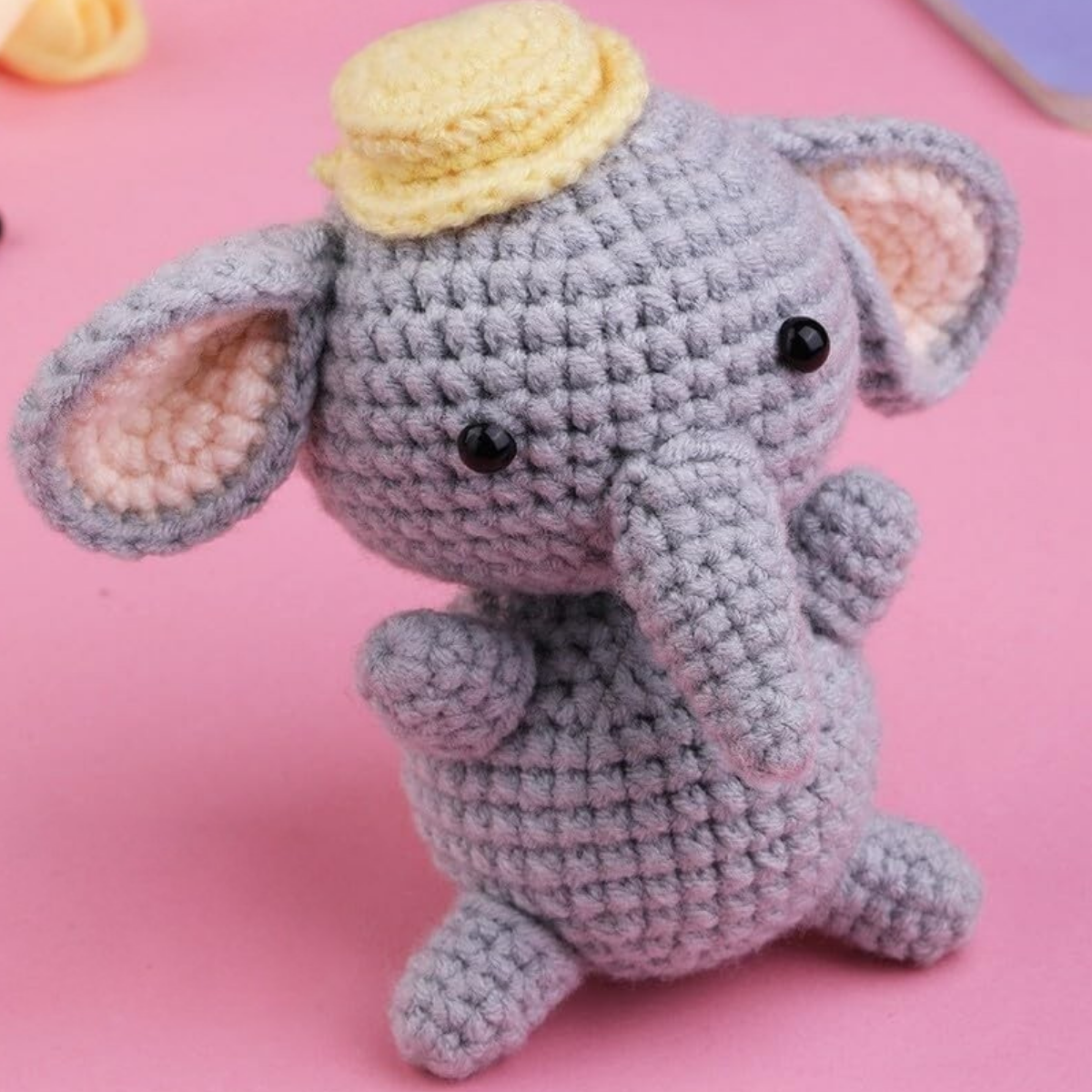 A gray elephant plush made with the Crochet Elephant Kit, featuring black eyes and a small yellow hat, sits on a pink surface.
