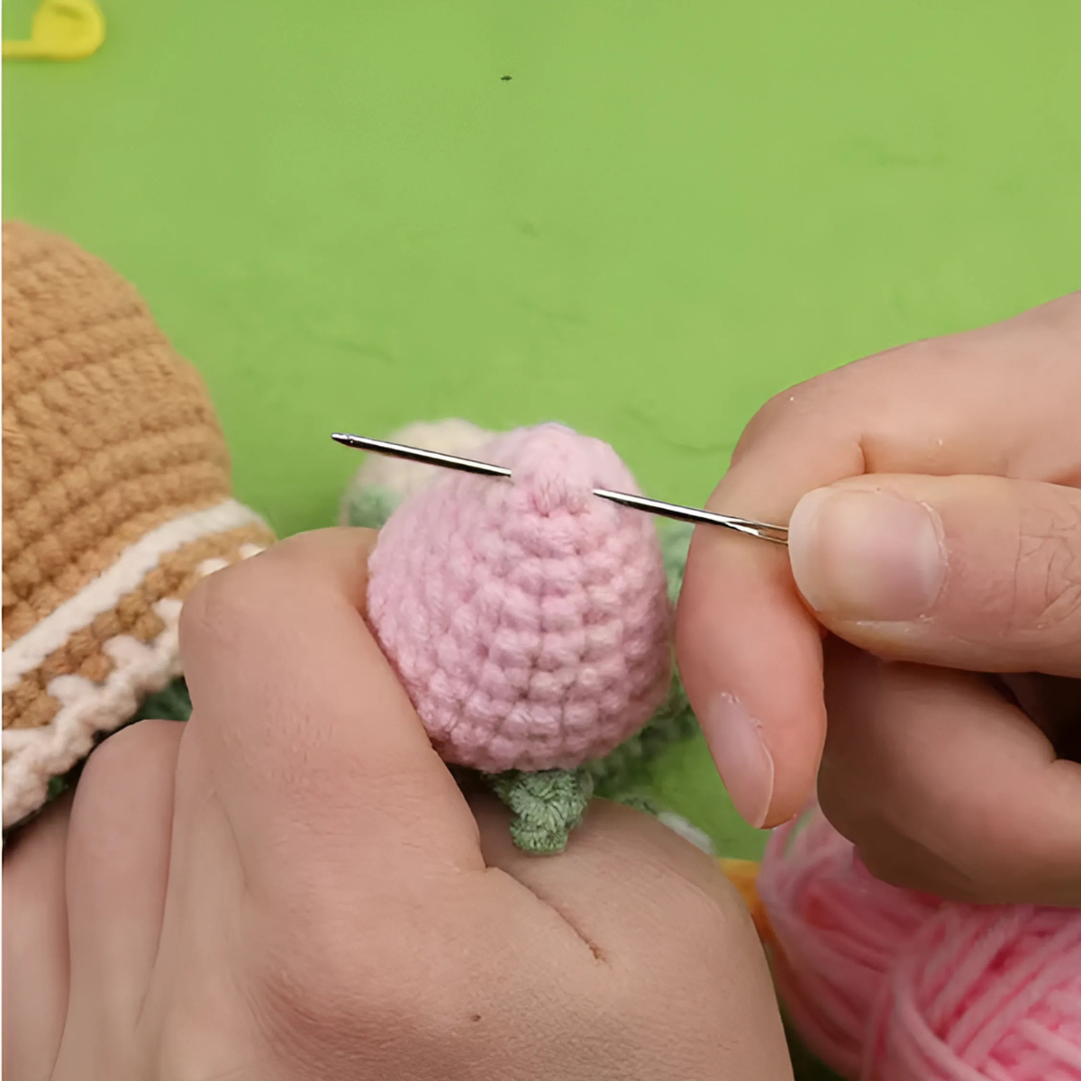 A person sews a pink piece from the Crochet Tulip Kit with a needle, holding it in one hand against a green background.