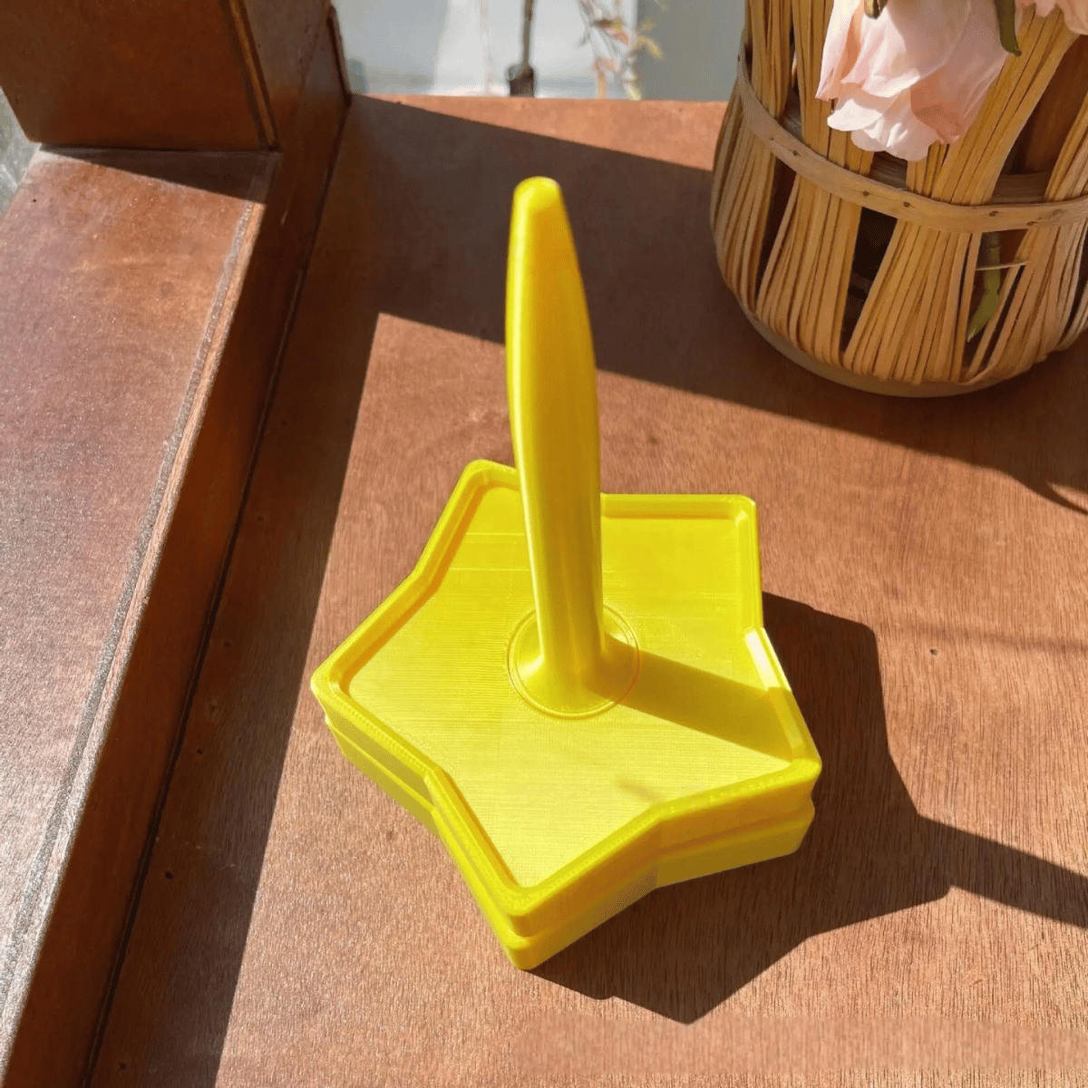 A Spinning Yarn Holder, featuring a yellow plastic star-shaped base and vertical handle, sits on a wooden surface beside a woven basket of flowers.