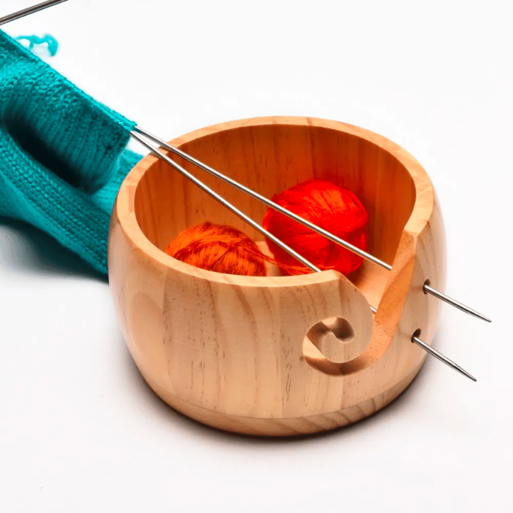 The Wooden Yarn Bowl With Lid cradles two orange yarn balls and serves as an ideal knitting accessory. Knitting needles with a turquoise yarn project rest on the bowl's edge, highlighting its elegance and functionality as a yarn dispenser.