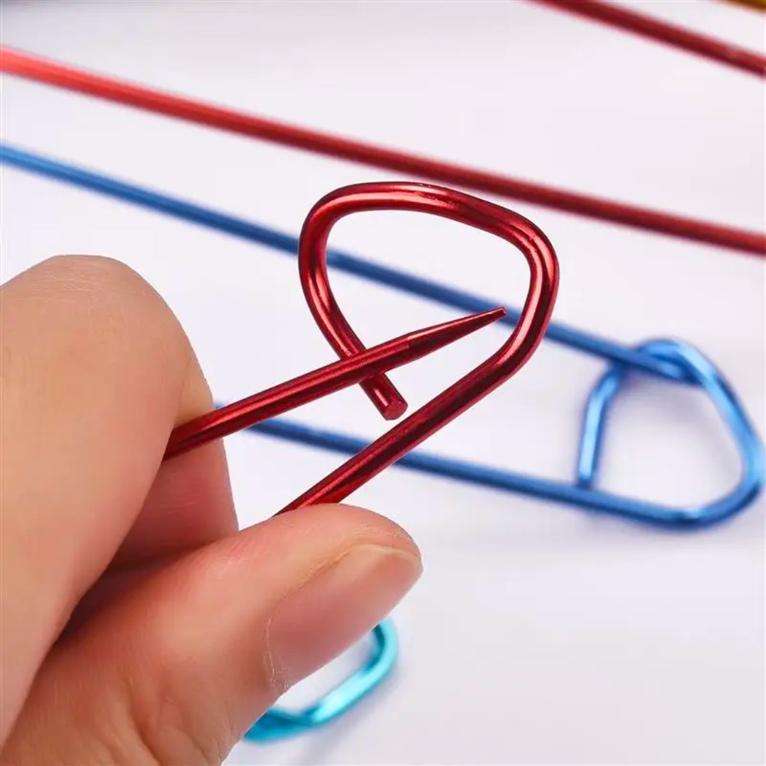 A close-up of a hand holding a red heart-shaped paperclip evokes the image of the vibrant tools from the Stitch Holder Set 6pcs. Nearby, several colorful and durable aluminum paperclips lay on the light background, resembling a stitch holder set ready for use.