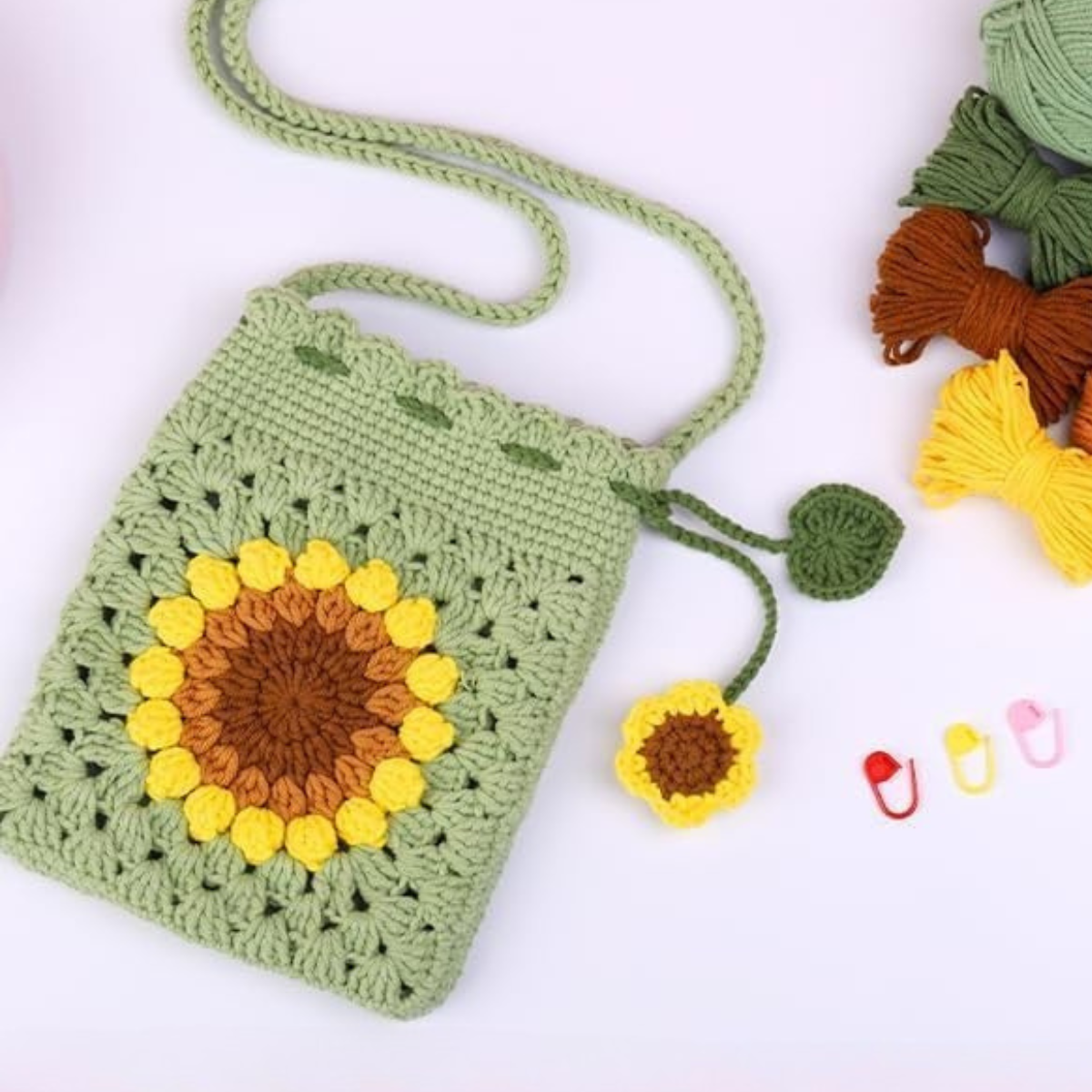 The Crochet Bag Kit features a green crocheted sunflower bag, a small sunflower charm, yarn skeins, and plastic stitch markers—ideal for any crochet enthusiast.
