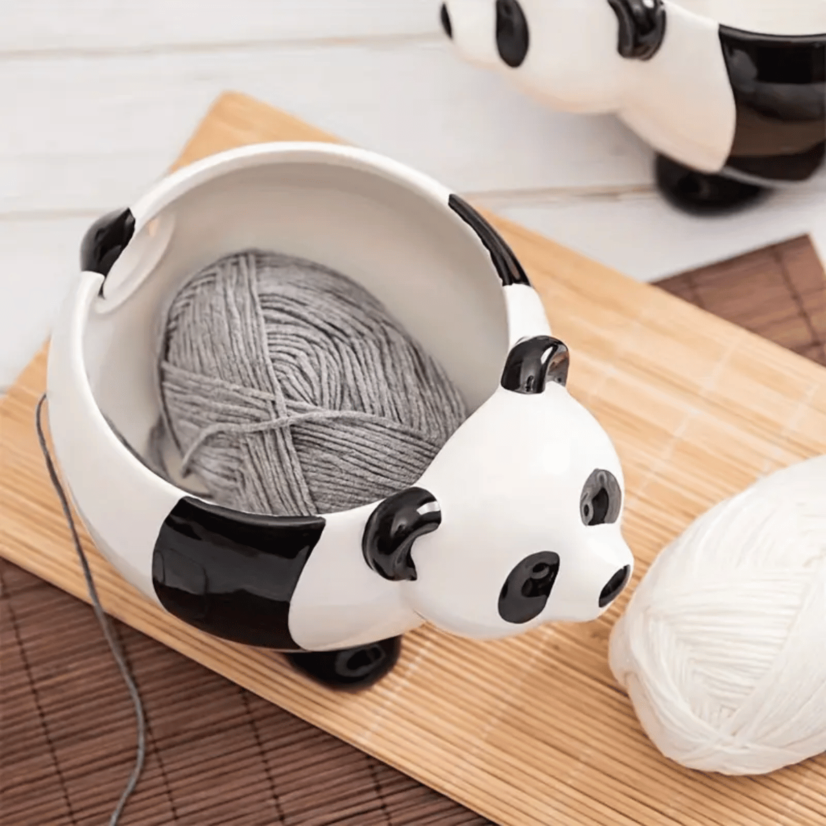 The Panda Ceramic Yarn Bowl holds a gray yarn ball with a strand threading out, while a white yarn ball sits beside it on a bamboo mat—ideal for beginner-friendly projects like cozy blankets or pairing with your favorite knitting loom.