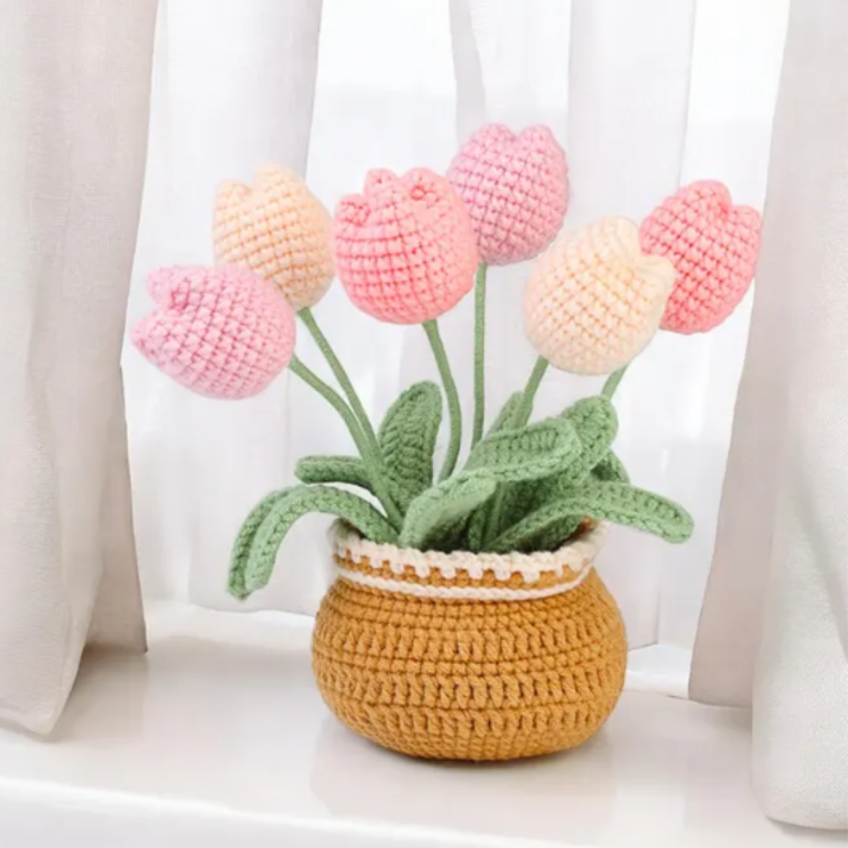 A Crochet Tulip Kit creates pink and peach tulips with green leaves, arranged in a yellow-brown basket and displayed on a windowsill before white curtains.