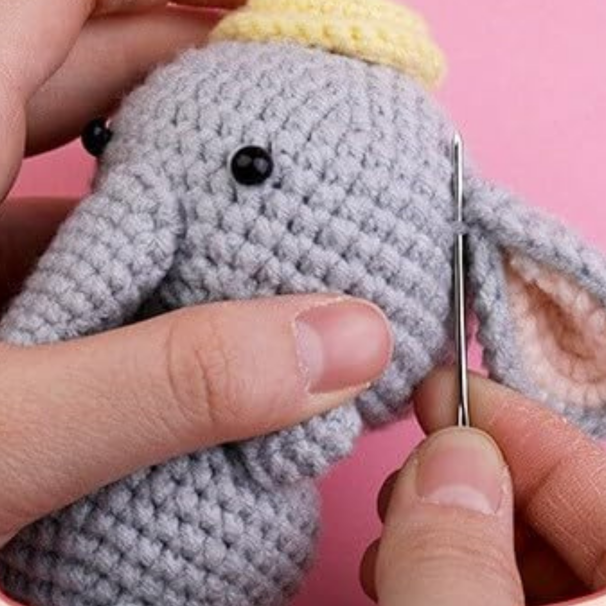 Two hands use the Crochet Elephant Kit to sew an elephant with a yellow hat, using yarn and a needle, set against a pink background.