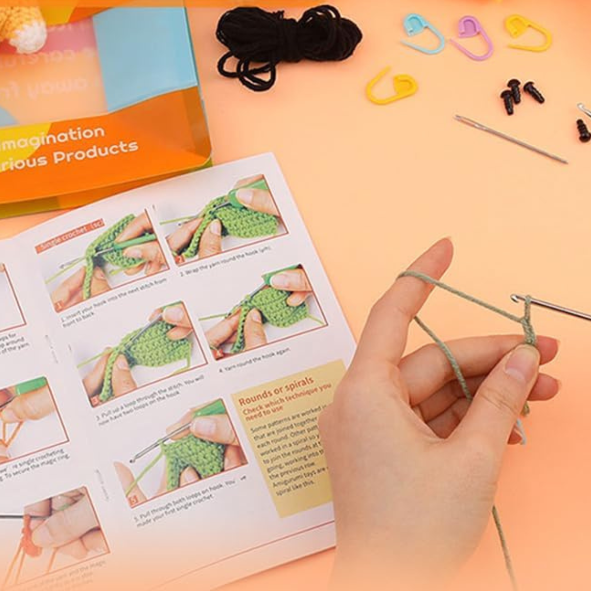 A hand begins crocheting with green yarn and a hook from the Crochet Bear Kit, alongside an instruction booklet, stitch markers, a needle, black yarn, and small accessories on a peach surface.