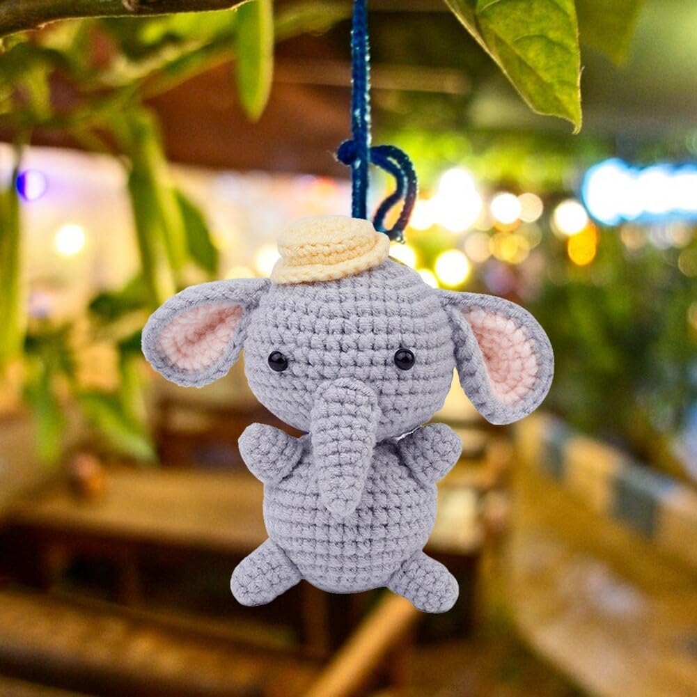 A gray elephant keychain, crafted with the Crochet Elephant Kit and wearing a small hat, hangs from a blue string on a branch against a softly blurred outdoor background.