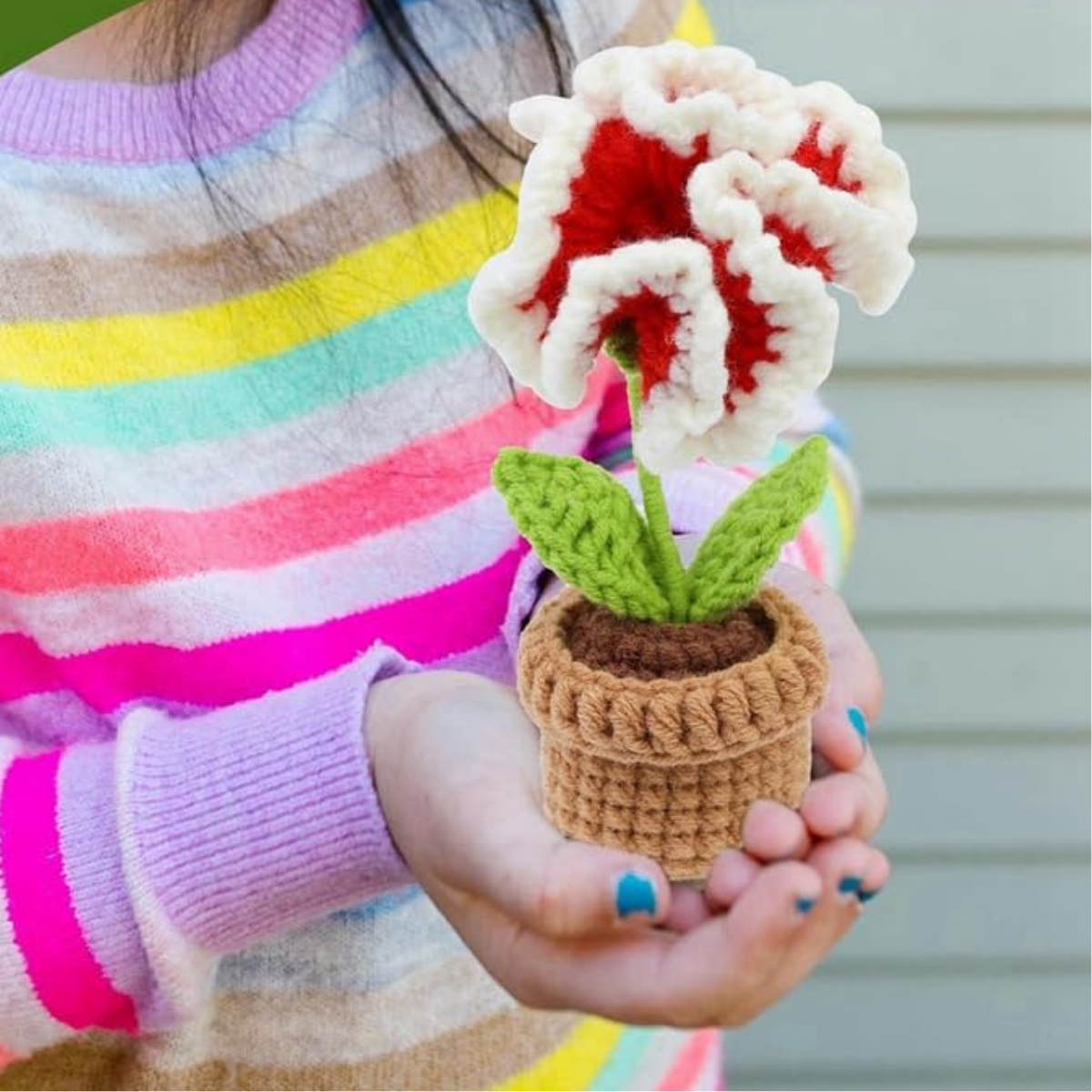 A person holds a handmade crochet flower from the Crochet Flower Pot Kit 6pcs, featuring red and white petals with green leaves in a small brown pot.