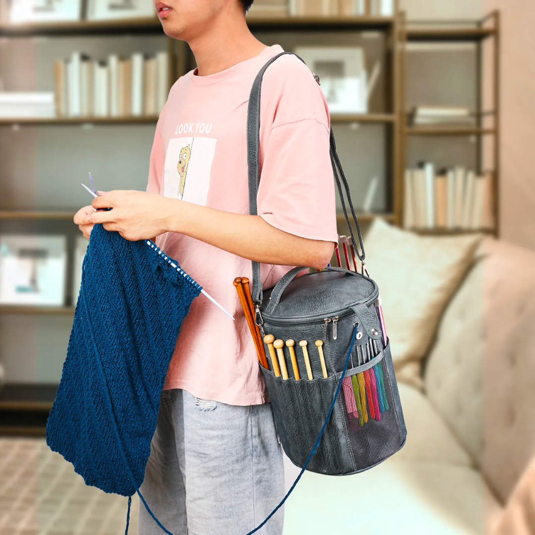 A person is knitting with blue yarn, their PU Leather Knitting Crochet Tools Storage Bag brimming with knitting needles and supplies. Shelves filled with books provide a cozy backdrop.