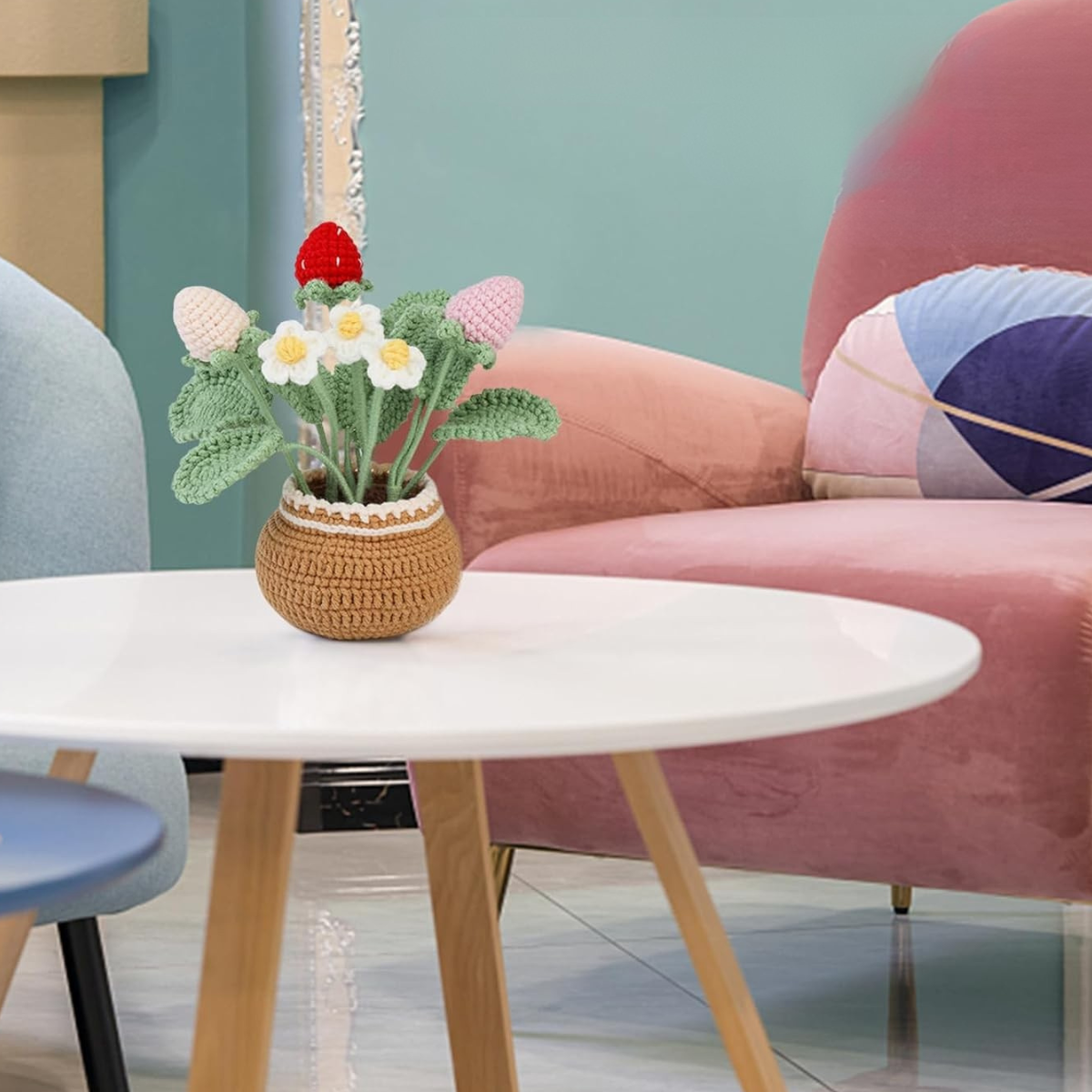 A small brown pot made from the Crochet Strawberry Plant Kit, filled with handmade strawberries, sits on a round white coffee table in a modern living room with pastel furniture.