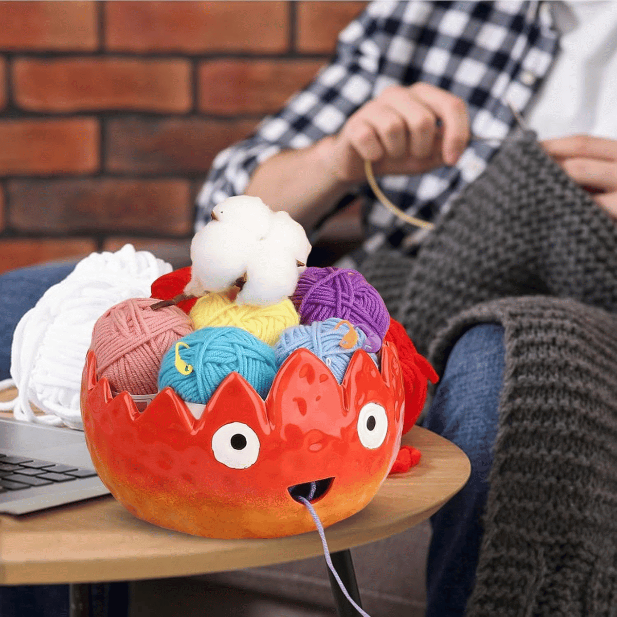 A person knits on a couch with a gray blanket; colorful yarn balls are in the Flame Ceramic Yarn Bowl, shaped like a red monster, on the coffee table—perfect for holding knit accessories.