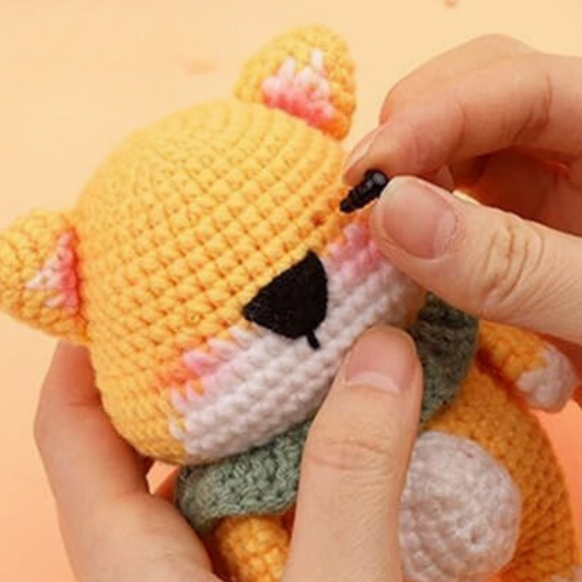 A person attaches a plastic eye to a crocheted yellow bear plush toy, made with the Crochet Bear Kit. The toy features a white snout, pink cheeks, and a green scarf.