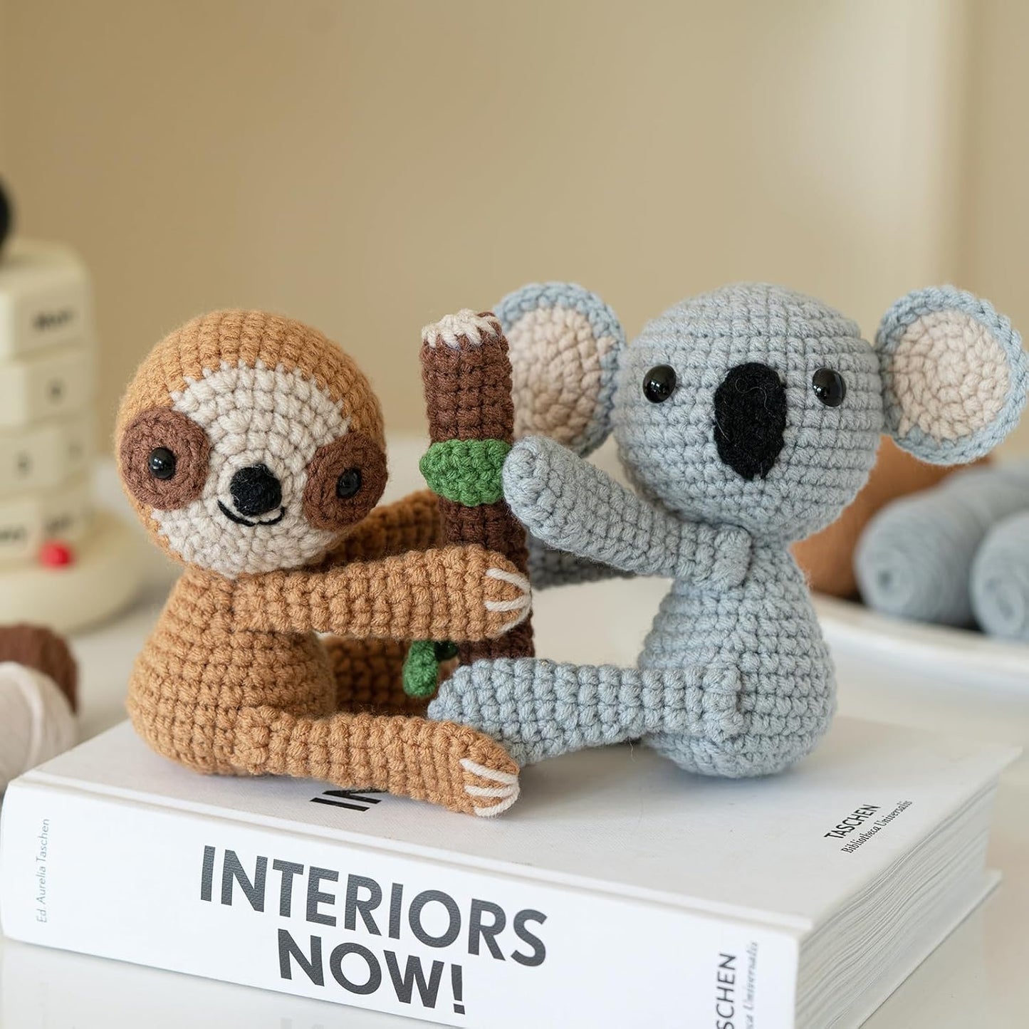 Two stuffed animals made with the Crochet Sloth & Koala Kit sit on a book called "INTERIORS NOW!" and hold a crocheted branch together.