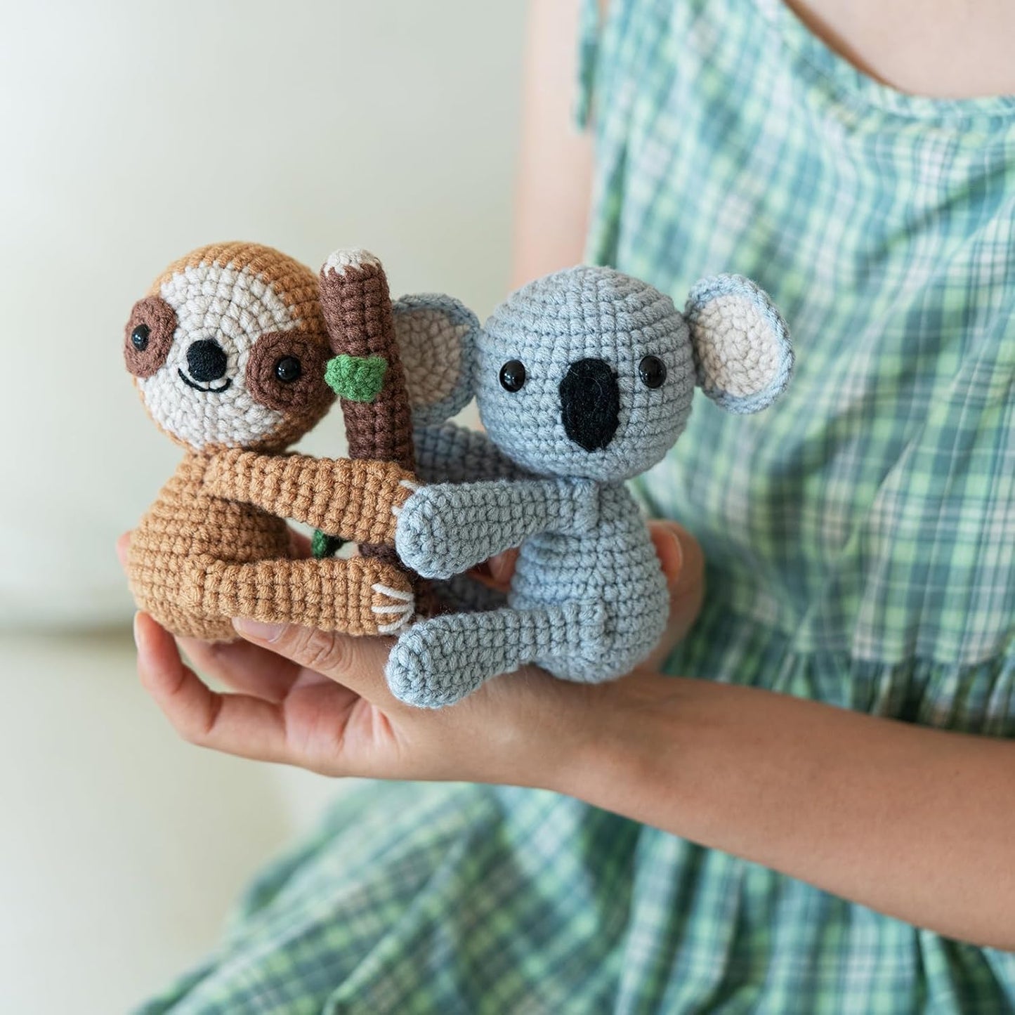 A person in a green checked dress holds two plush toys—a brown sloth and a gray koala made with the Crochet Sloth & Koala Kit—hugging a small crocheted branch.