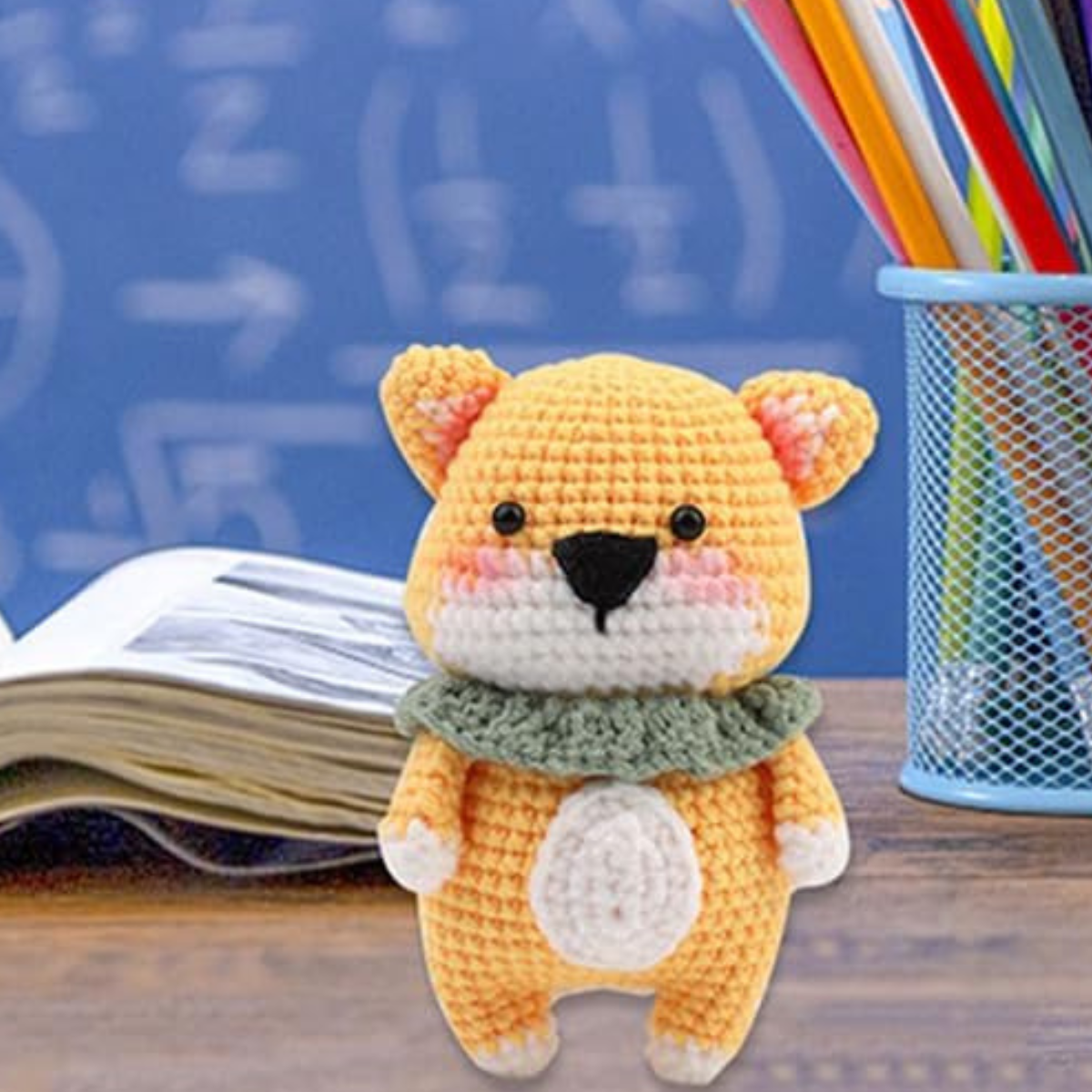 A bear made with the Crochet Bear Kit sits on a wooden desk in front of an open book and a pencil holder filled with colored pencils, with a chalkboard in the background.