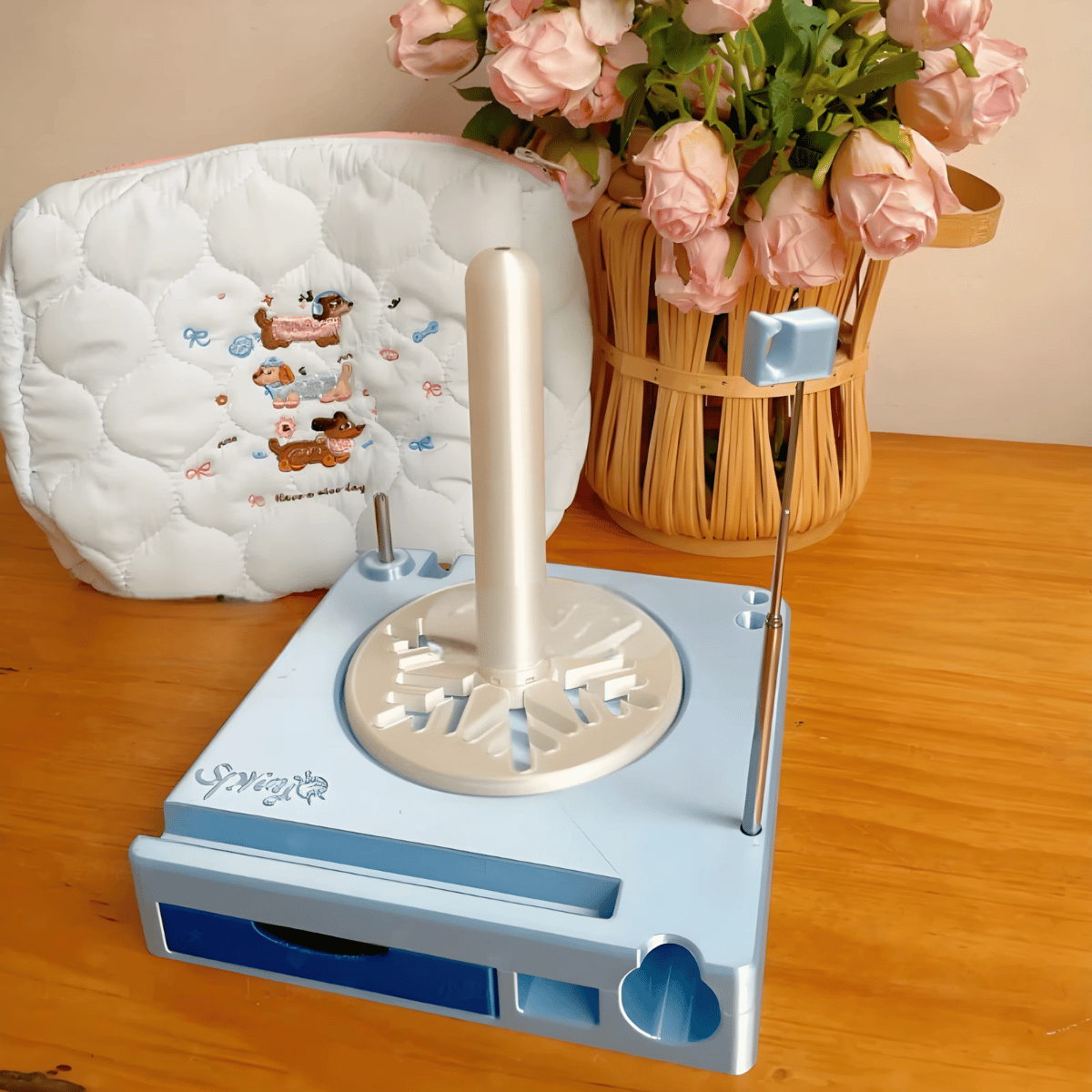 The Spinning Yarn Holder with Tool Storage, featuring a blue design, vertical spindle, and metal rods, sits on a wooden table next to pink roses and a white cushion, keeping your yarn flow smooth and organized.